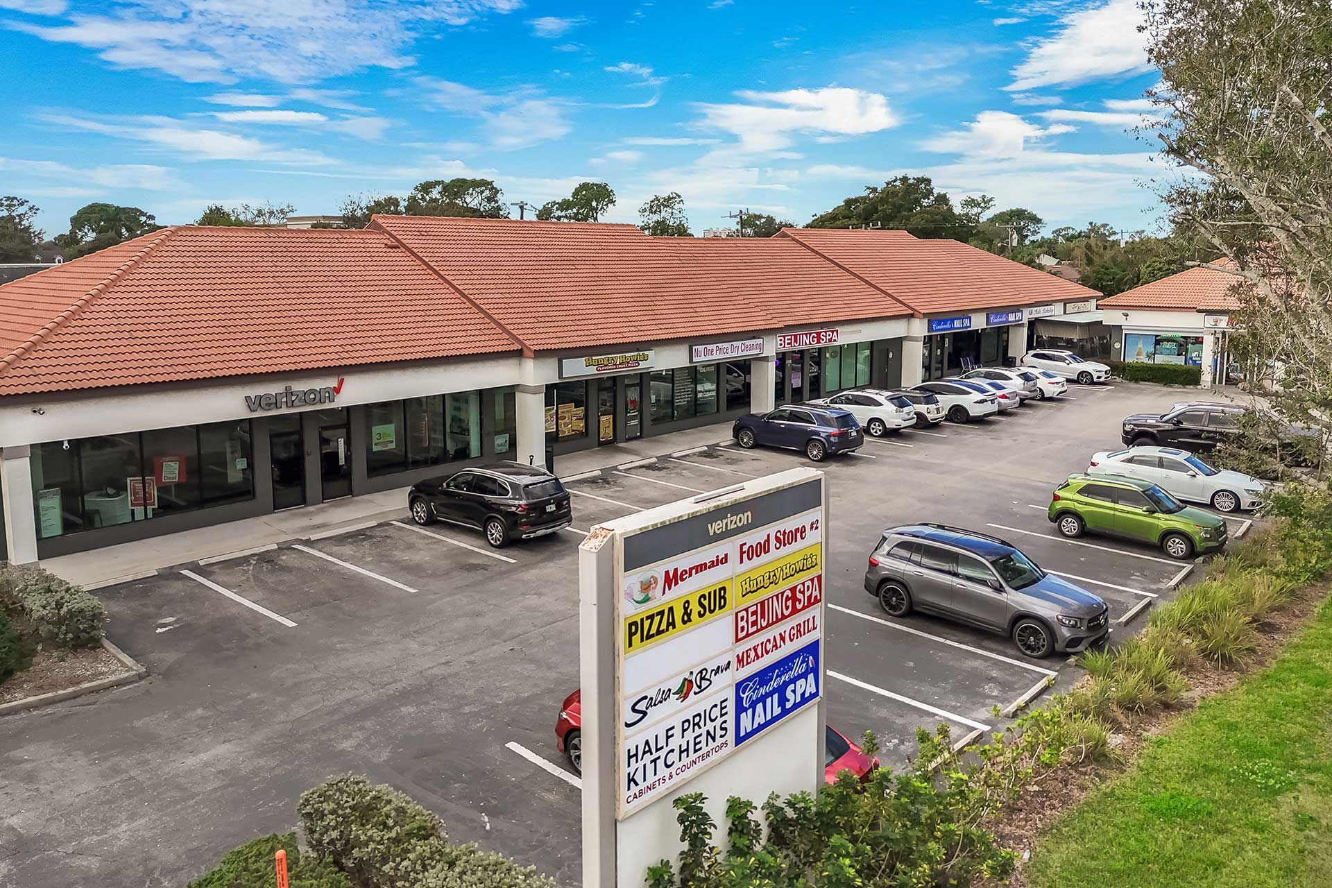 A shopping center with a red tile roof, stores, parked cars, and a sign. Blue sky.