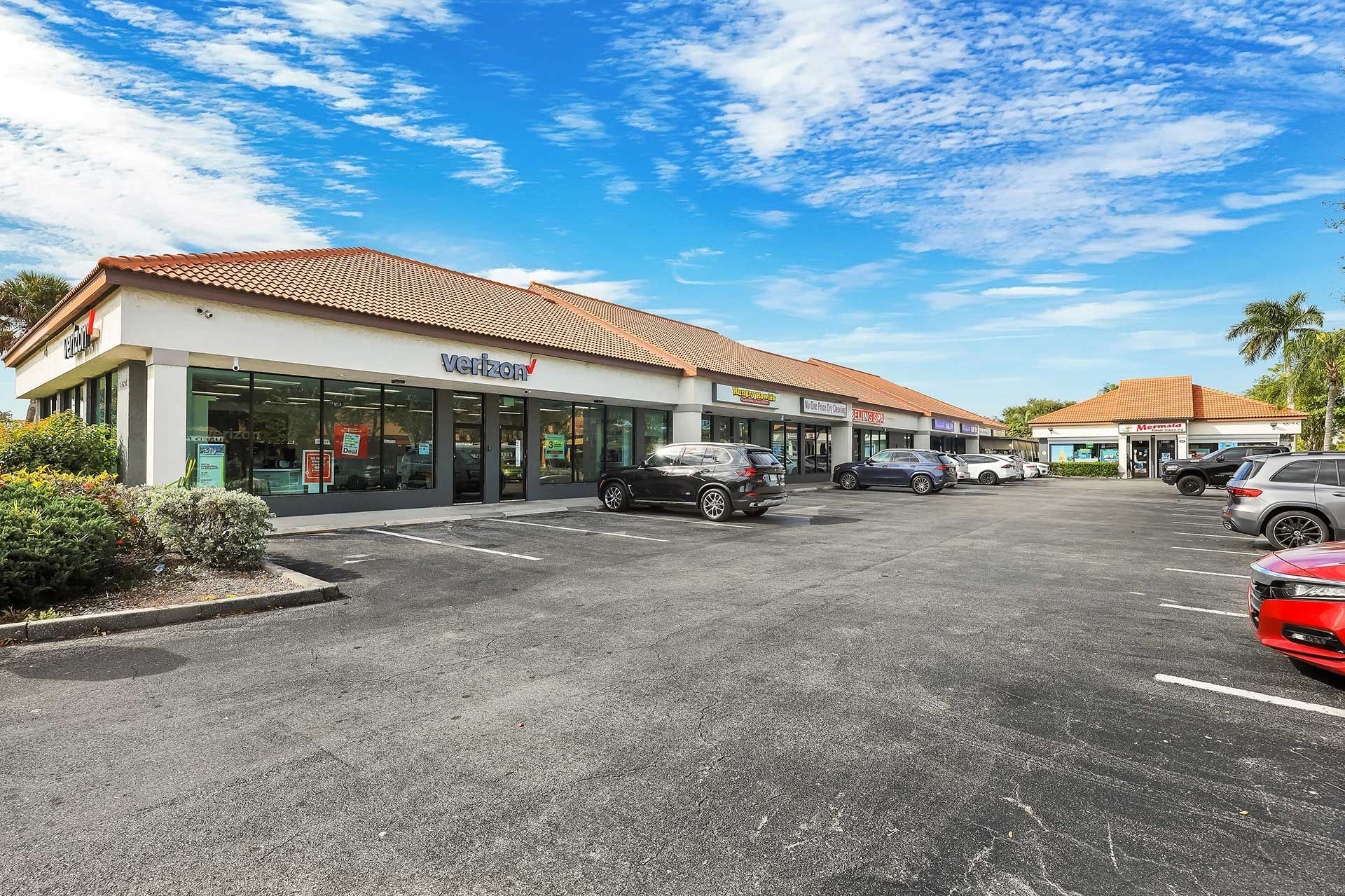 Commercial plaza with businesses, cars in parking lot, blue sky.