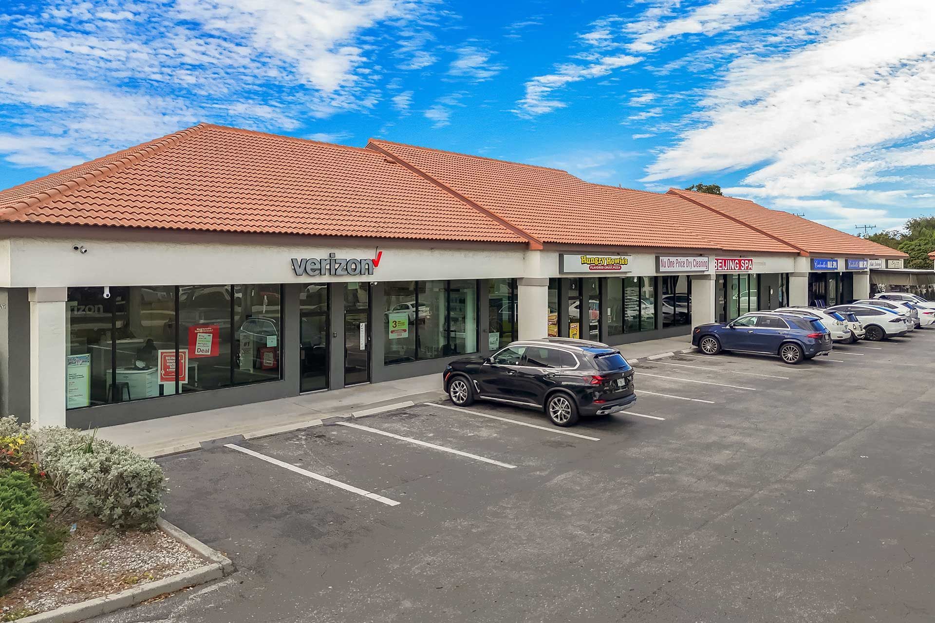 Shopping plaza with storefronts, parking, and cars against a blue sky.