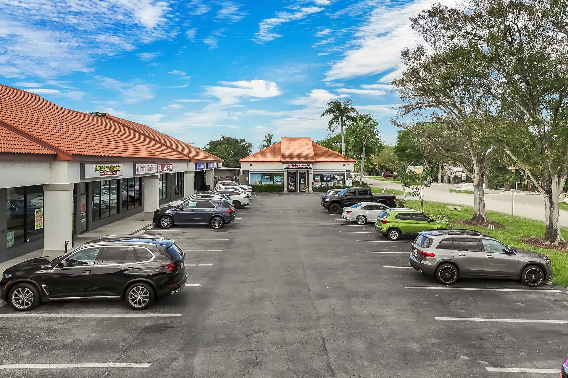 A strip mall with various storefronts, cars parked in front, and a blue sky overhead.