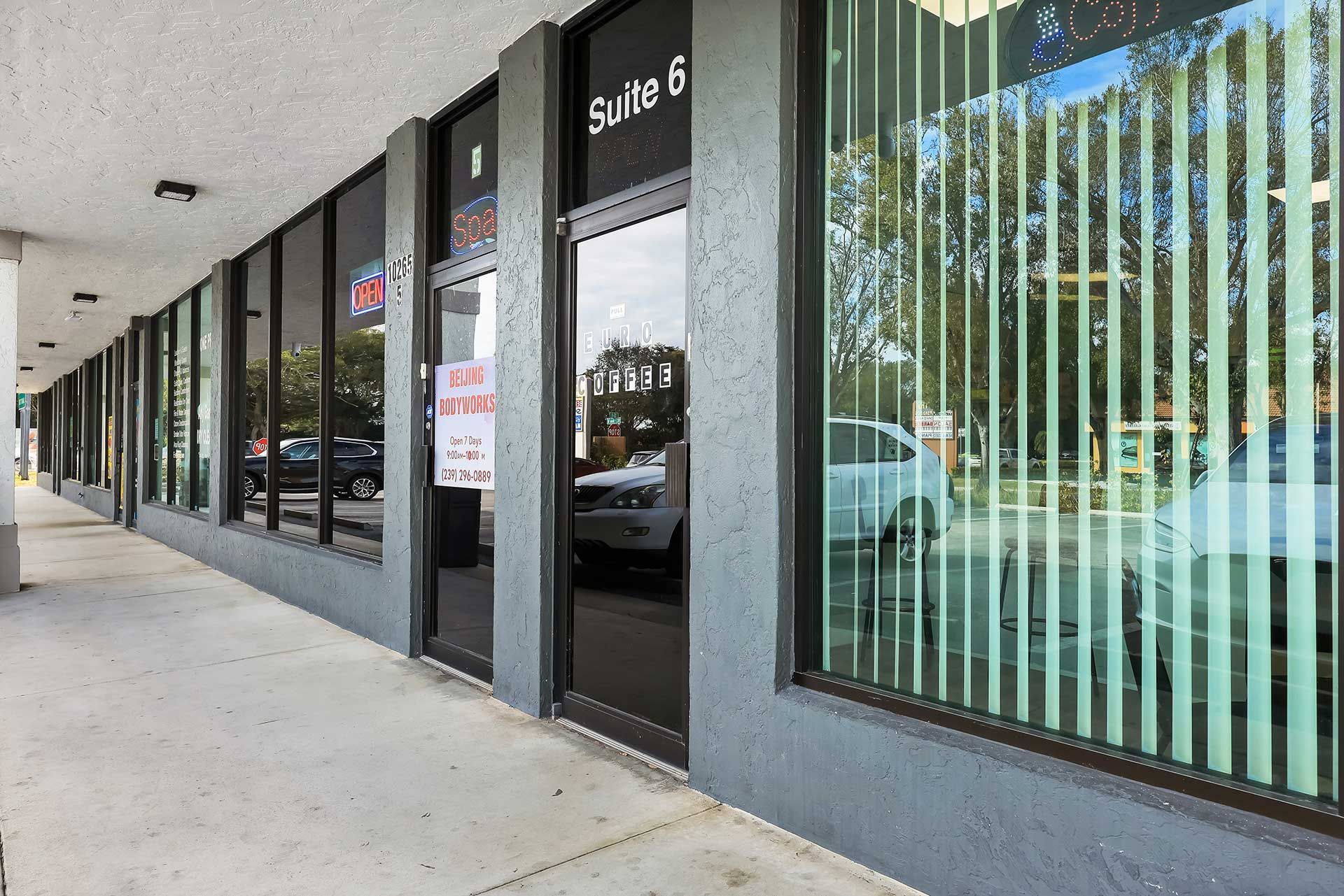 Exterior of a gray commercial building with glass windows, a sidewalk, and a sign that says Suite 6.