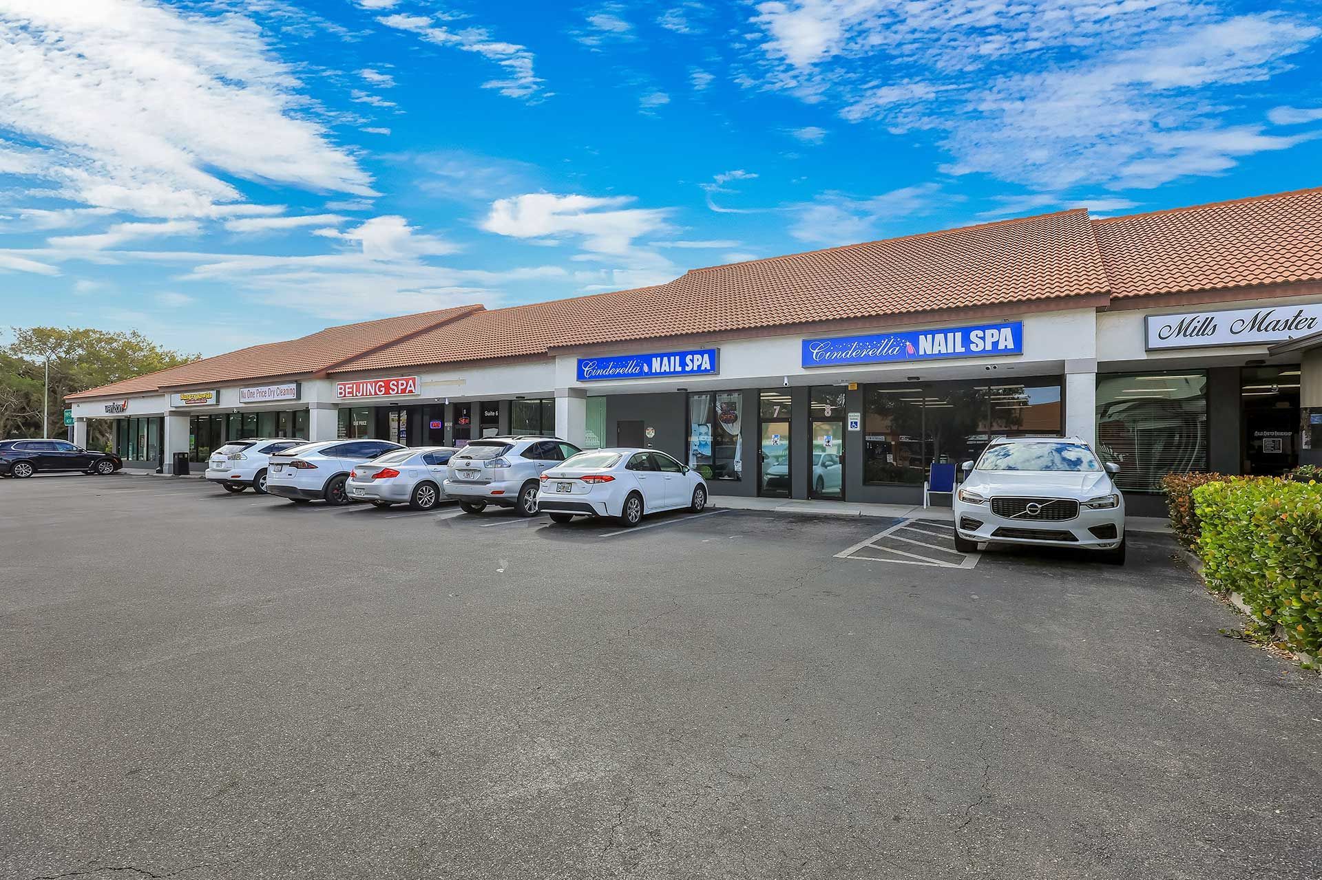 Strip mall with storefronts, parked cars, and blue sky with clouds.
