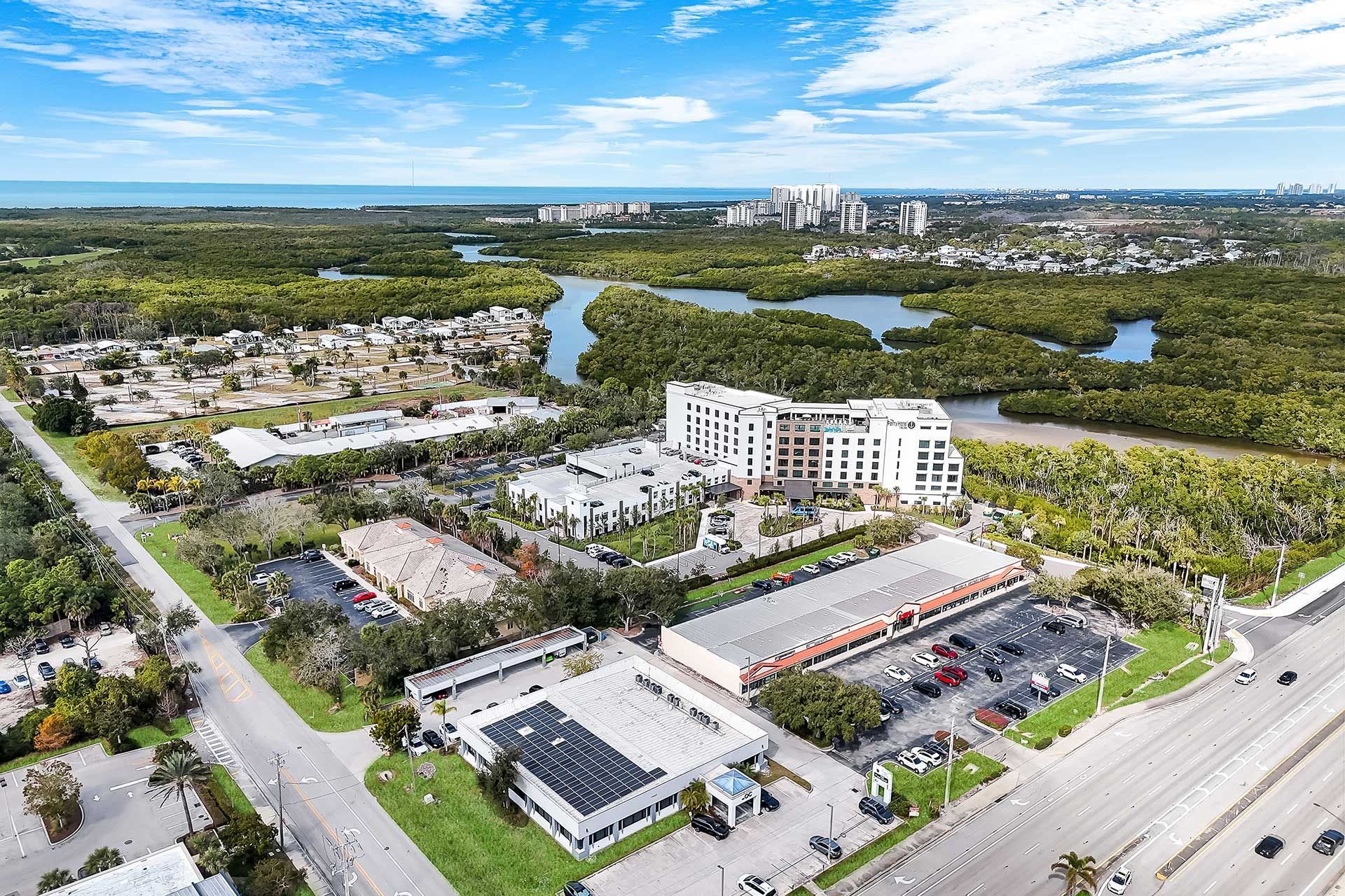 Aerial view of buildings, green trees, and water along a coastline under a blue sky.