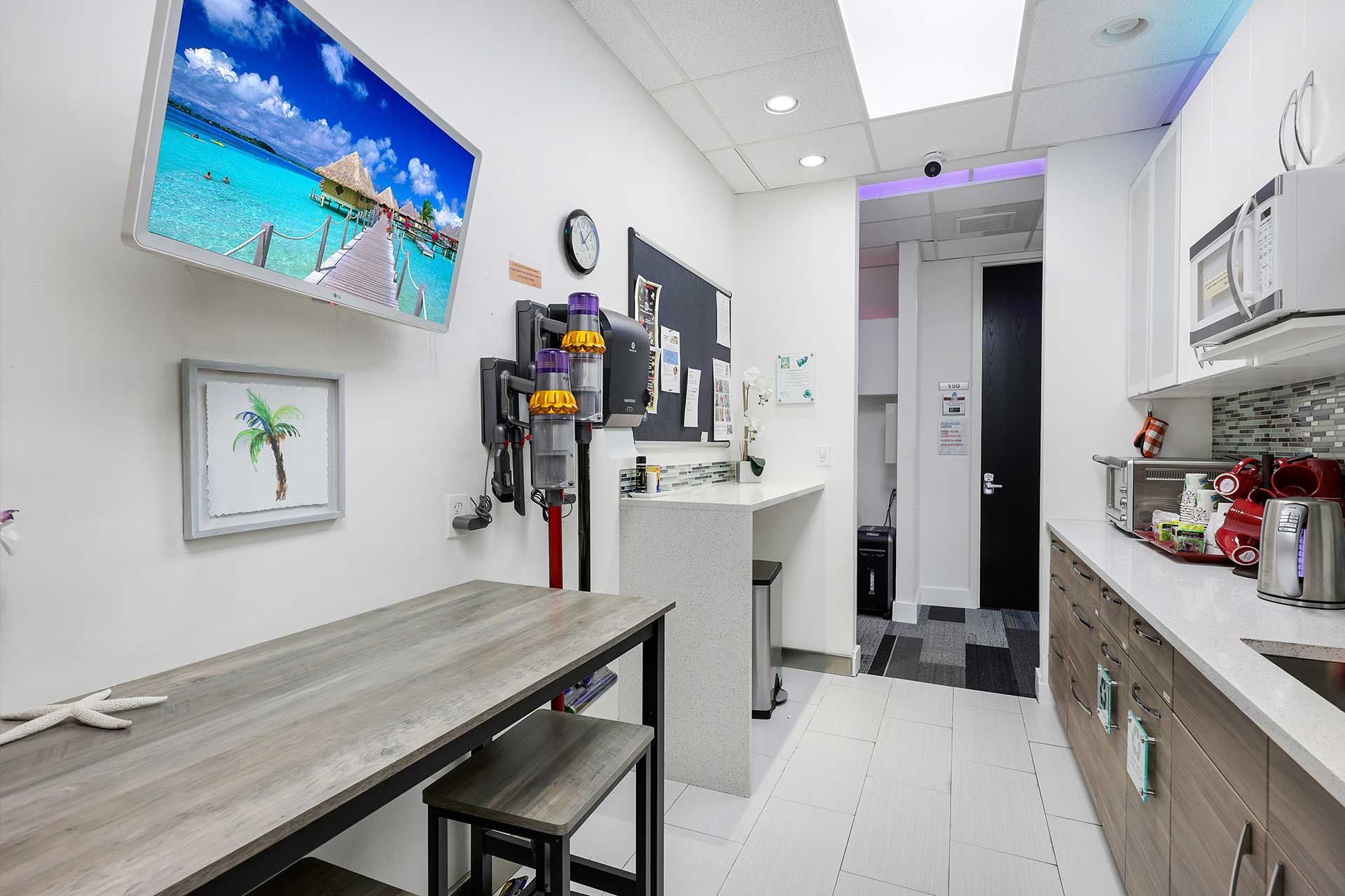 A brightly lit office kitchenette with a TV, vacuum, counter, and appliances.
