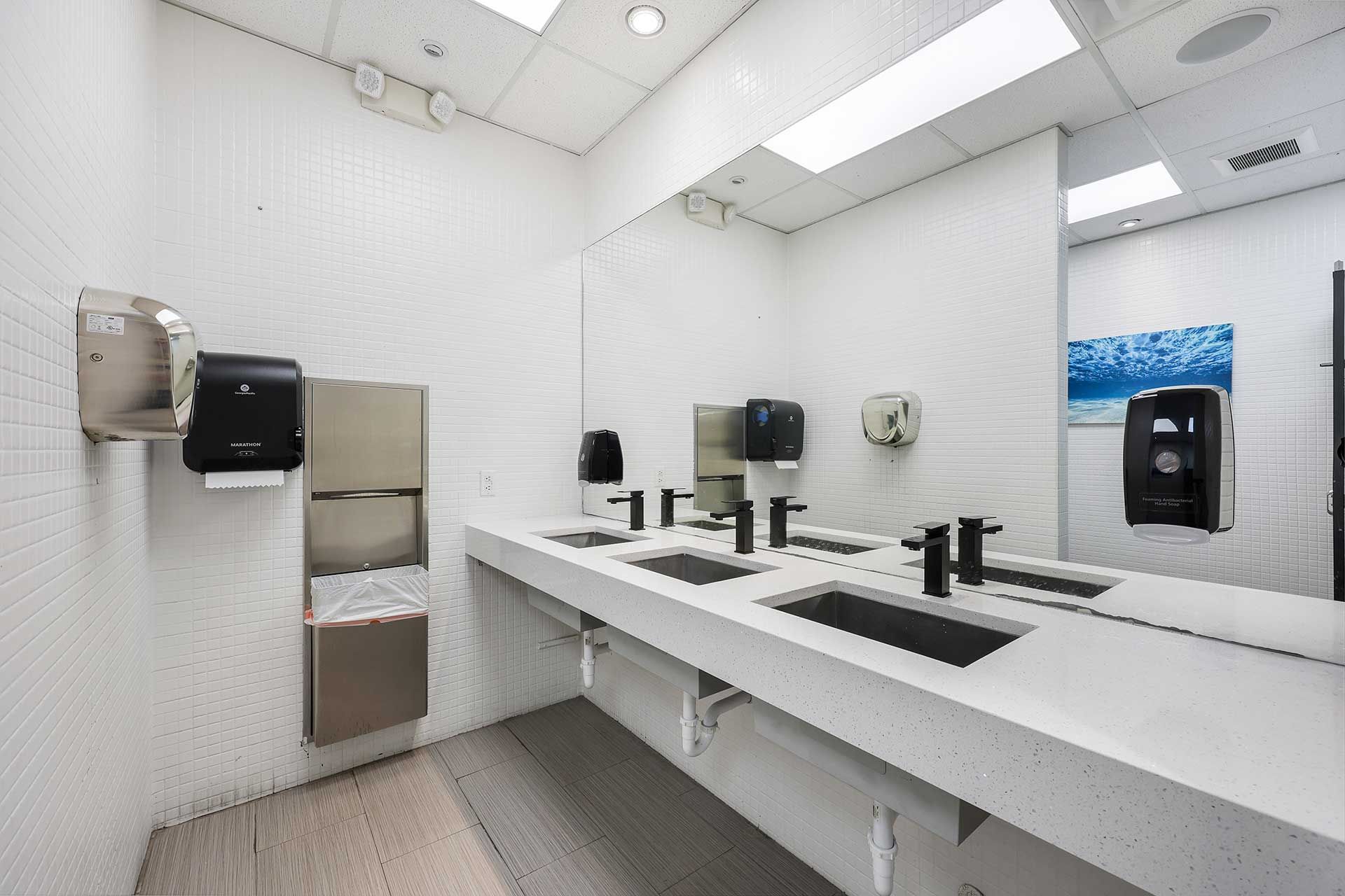 Modern, white tiled bathroom with long countertop, sinks, mirrors, and dispensers.