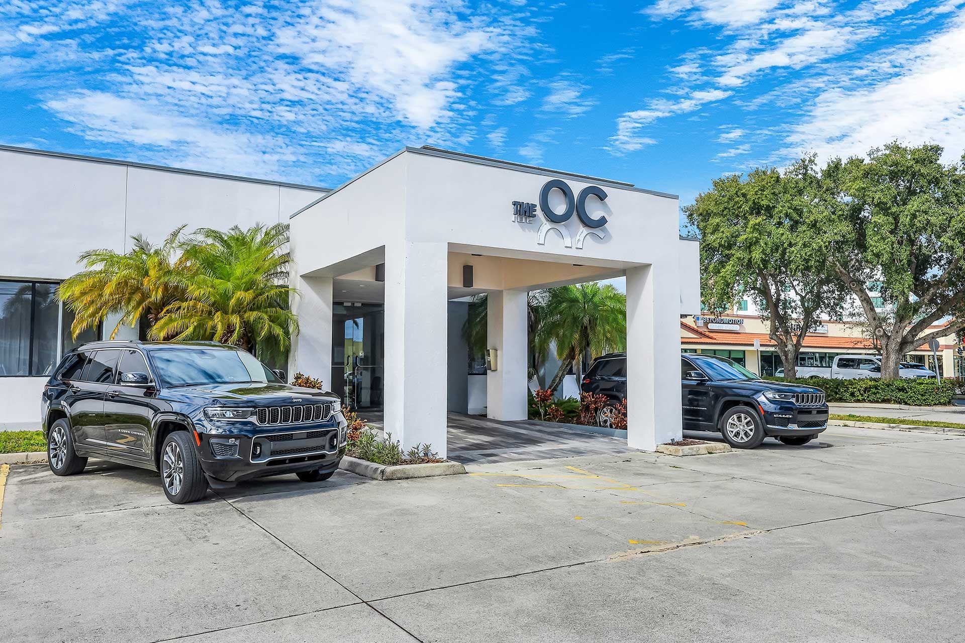 White building entrance with cars parked out front, under a blue sky.