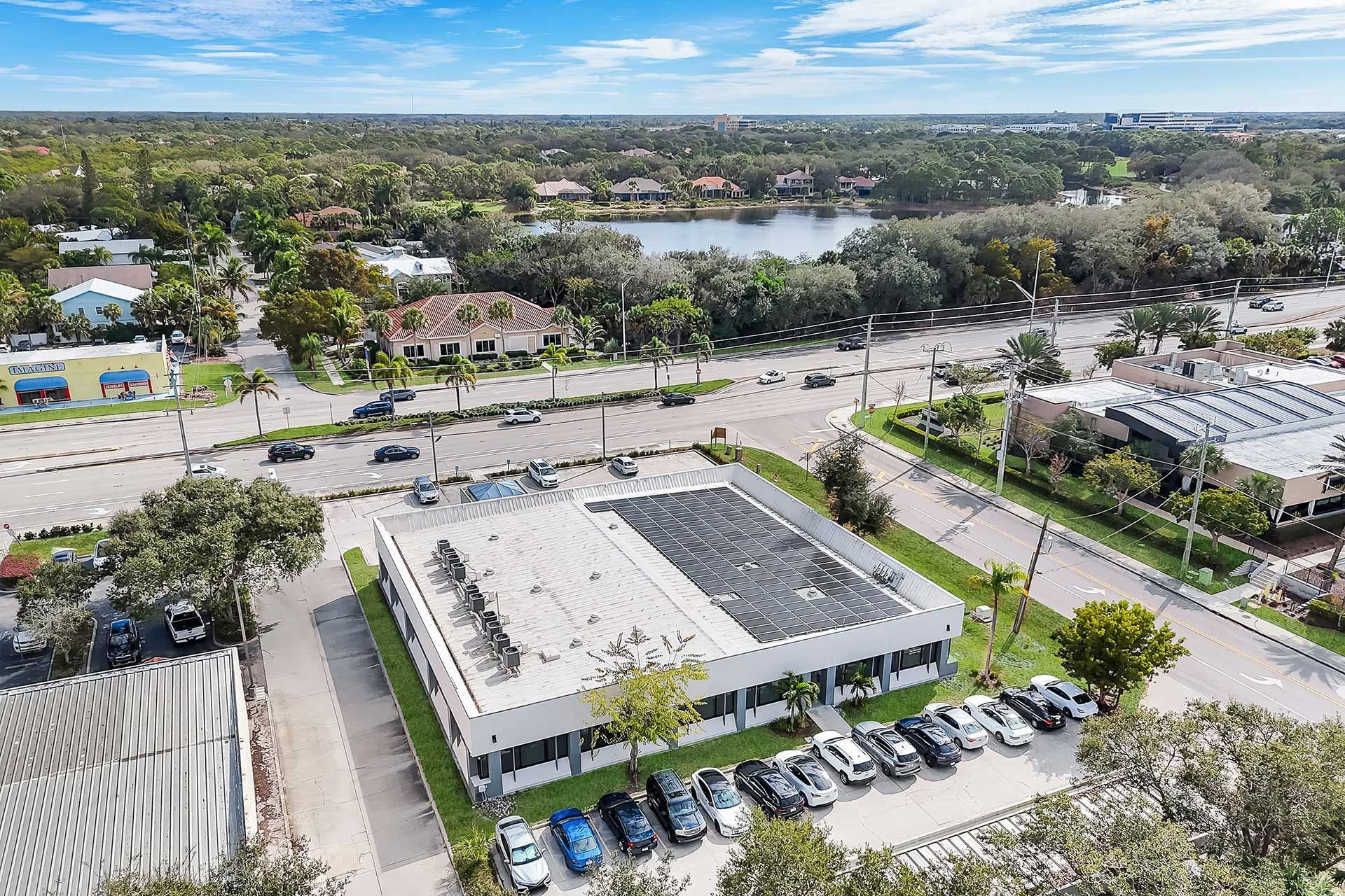 Aerial view of a white commercial building with cars parked in front, road, trees, and lake in background.