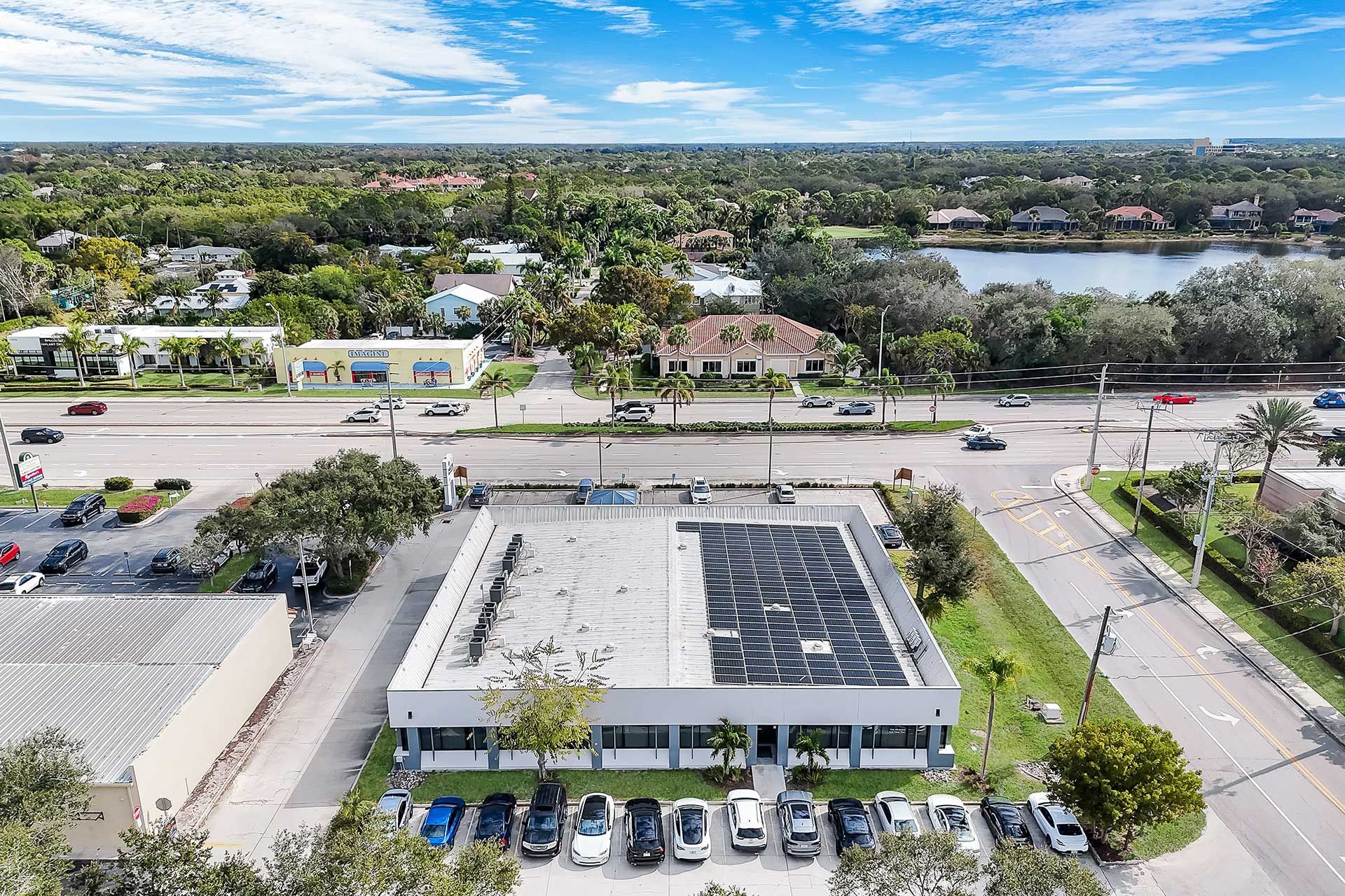 Aerial view of a gray building with parked cars, a road, and green trees under a blue sky.