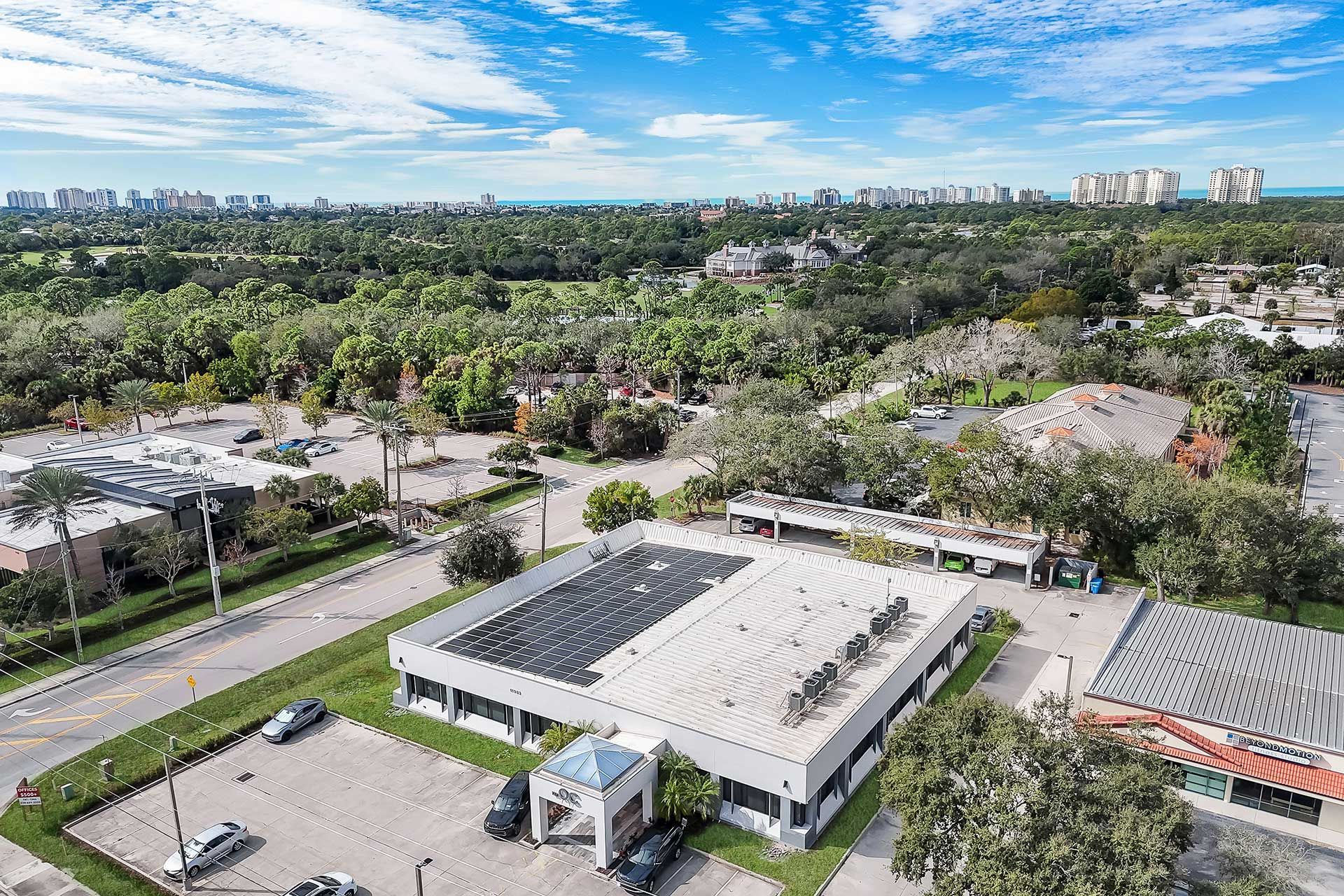Aerial view of a white, low-rise building with a flat roof; parking lot, trees, and city skyline in the background.