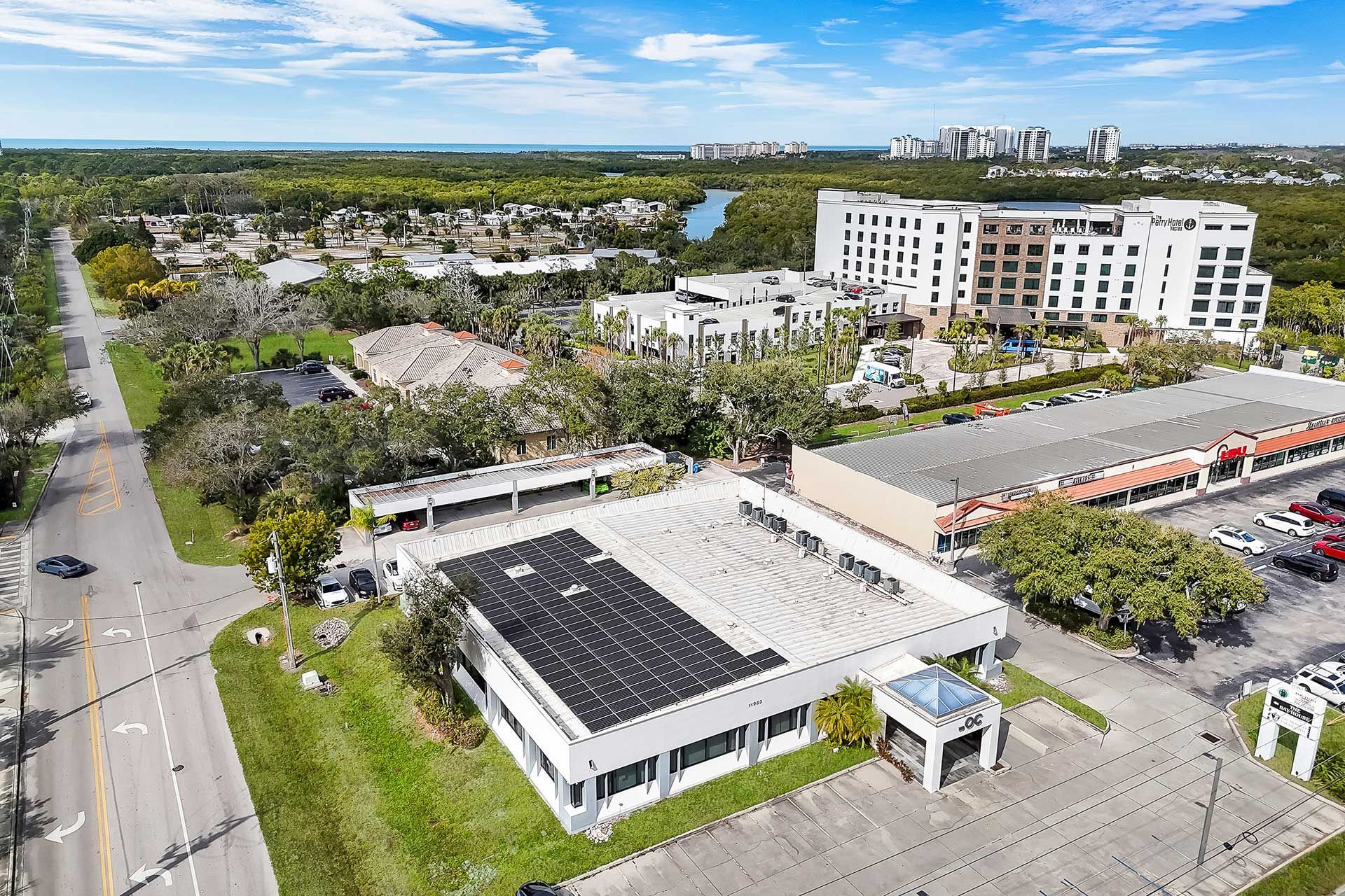 Aerial view of a commercial area with buildings, roads, trees, and a body of water in the distance.