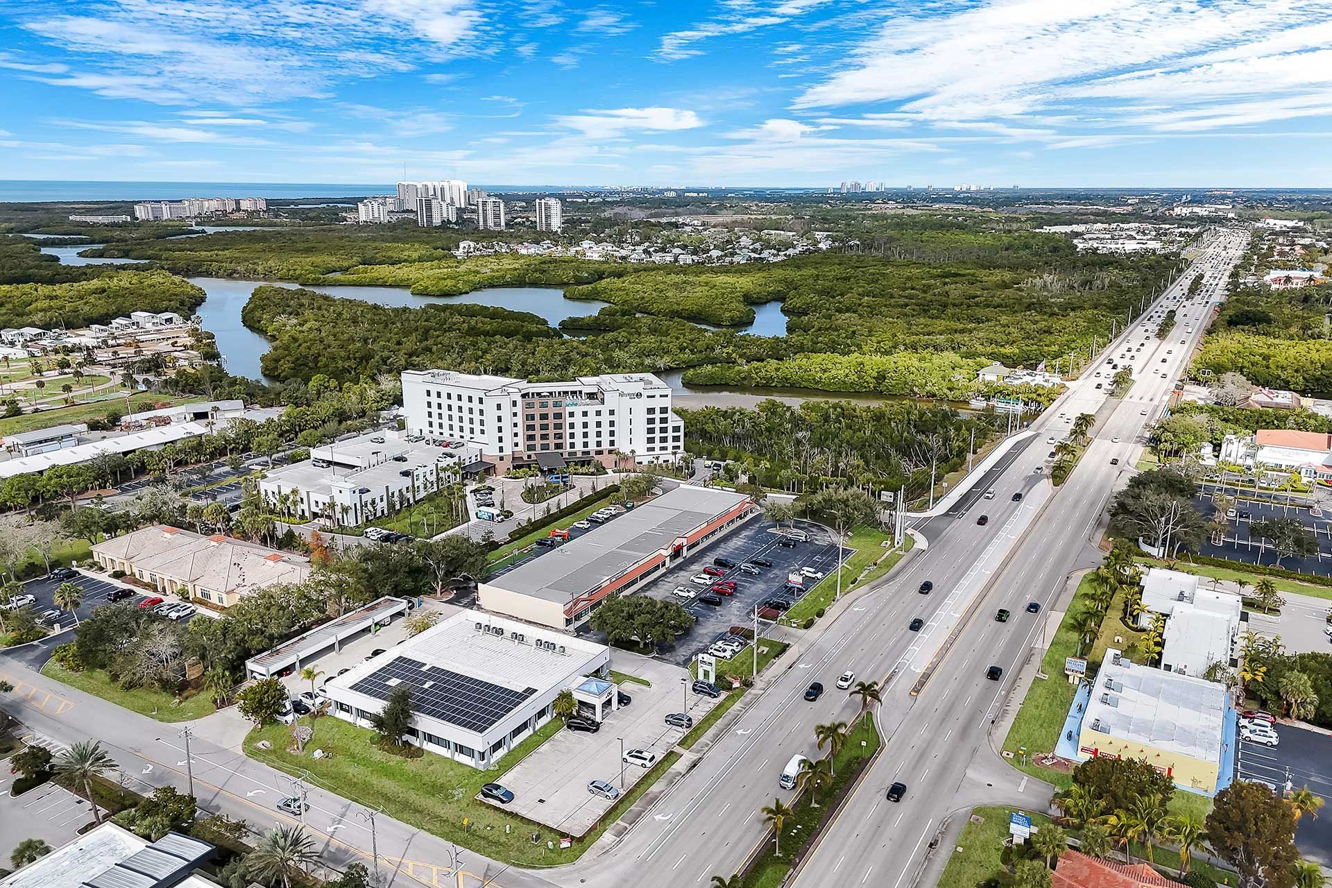 Aerial view of a coastal city with a wide road, buildings, and green wetlands under a blue sky.