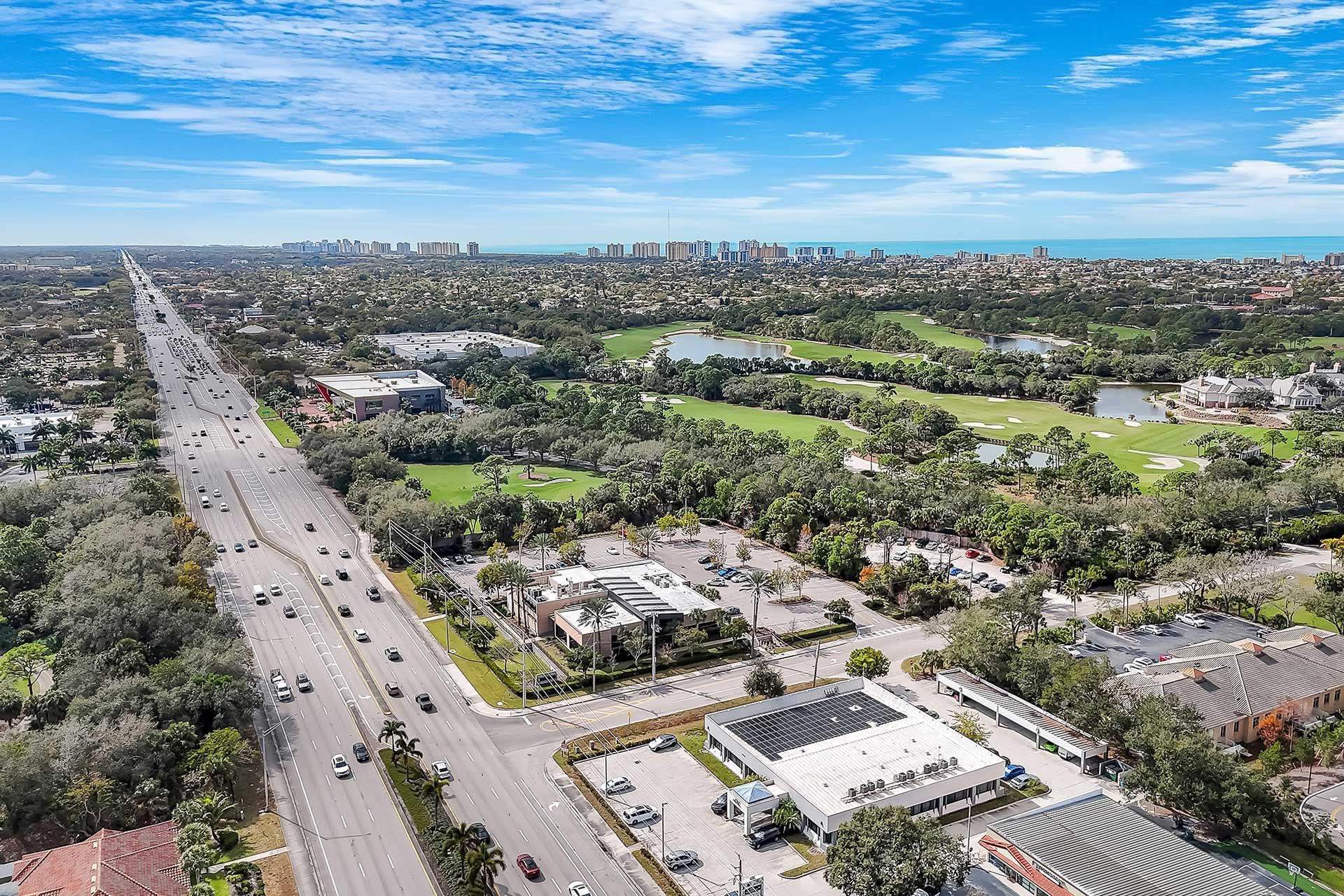 Aerial view: a wide road lined with trees, buildings, and cars, leading to the ocean under a blue sky.