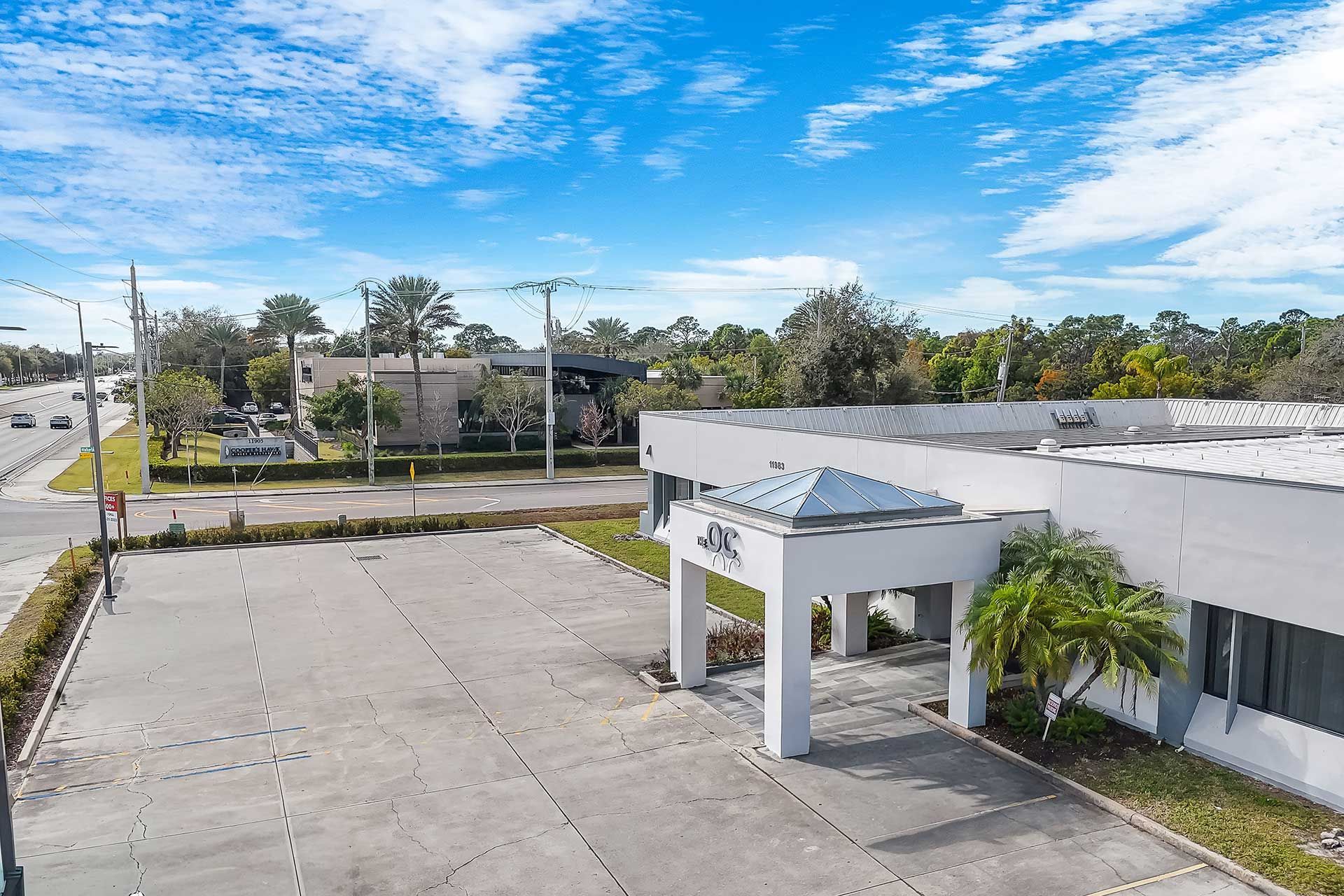 Building with angled roof, large parking lot, road, blue sky with clouds.