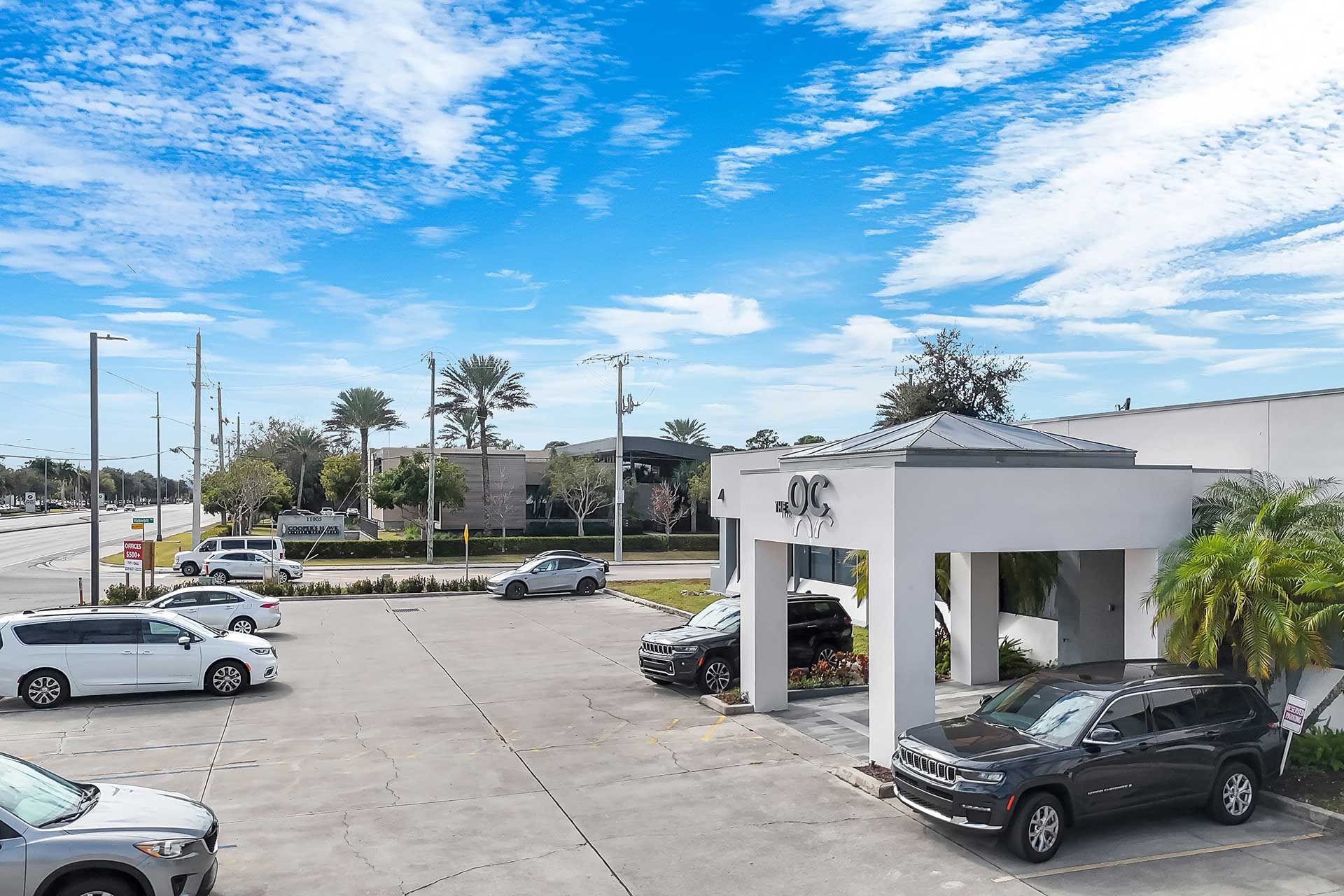 Vehicles parked in front of a building with a white entrance under a bright blue sky with wispy clouds.