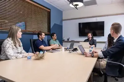 People at a table in a meeting, with laptop, TV screen, and neutral-toned wall.