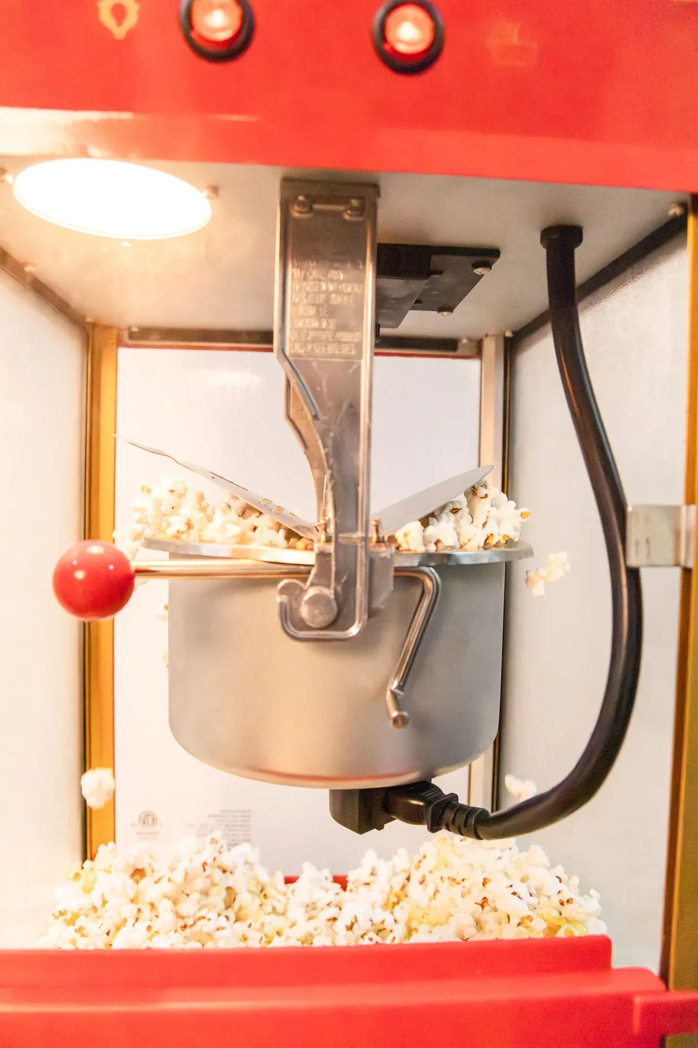 Popcorn machine: red exterior, silver kettle popping corn, kernels spilling into the base.