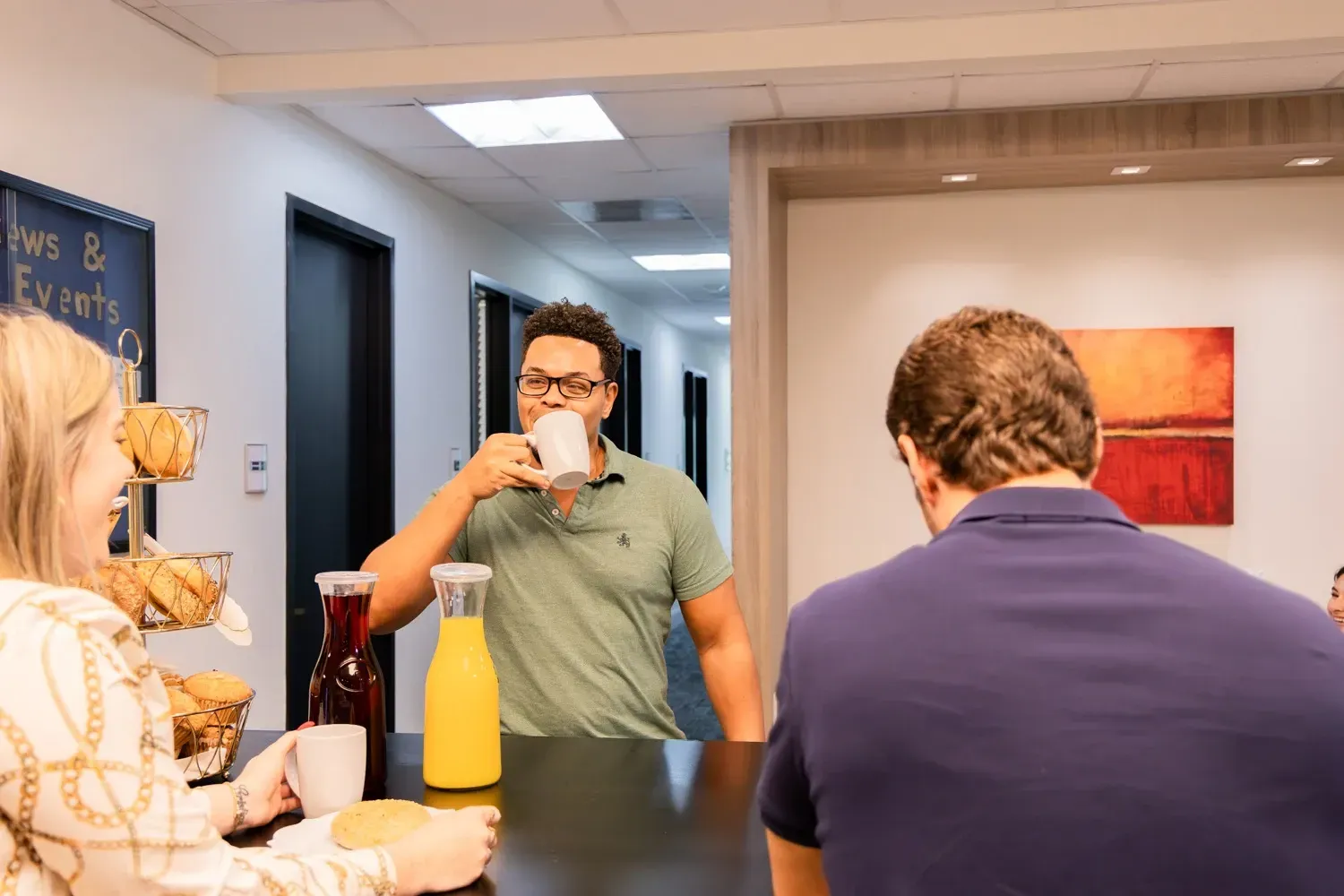 People in an office break room. One drinks from a mug, two others eat. Food and drinks are on the counter.