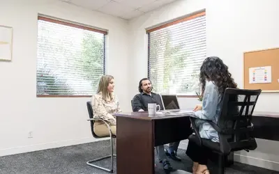 Three people in an office setting: a seated woman, a man, and another woman behind a desk, possibly in a meeting.