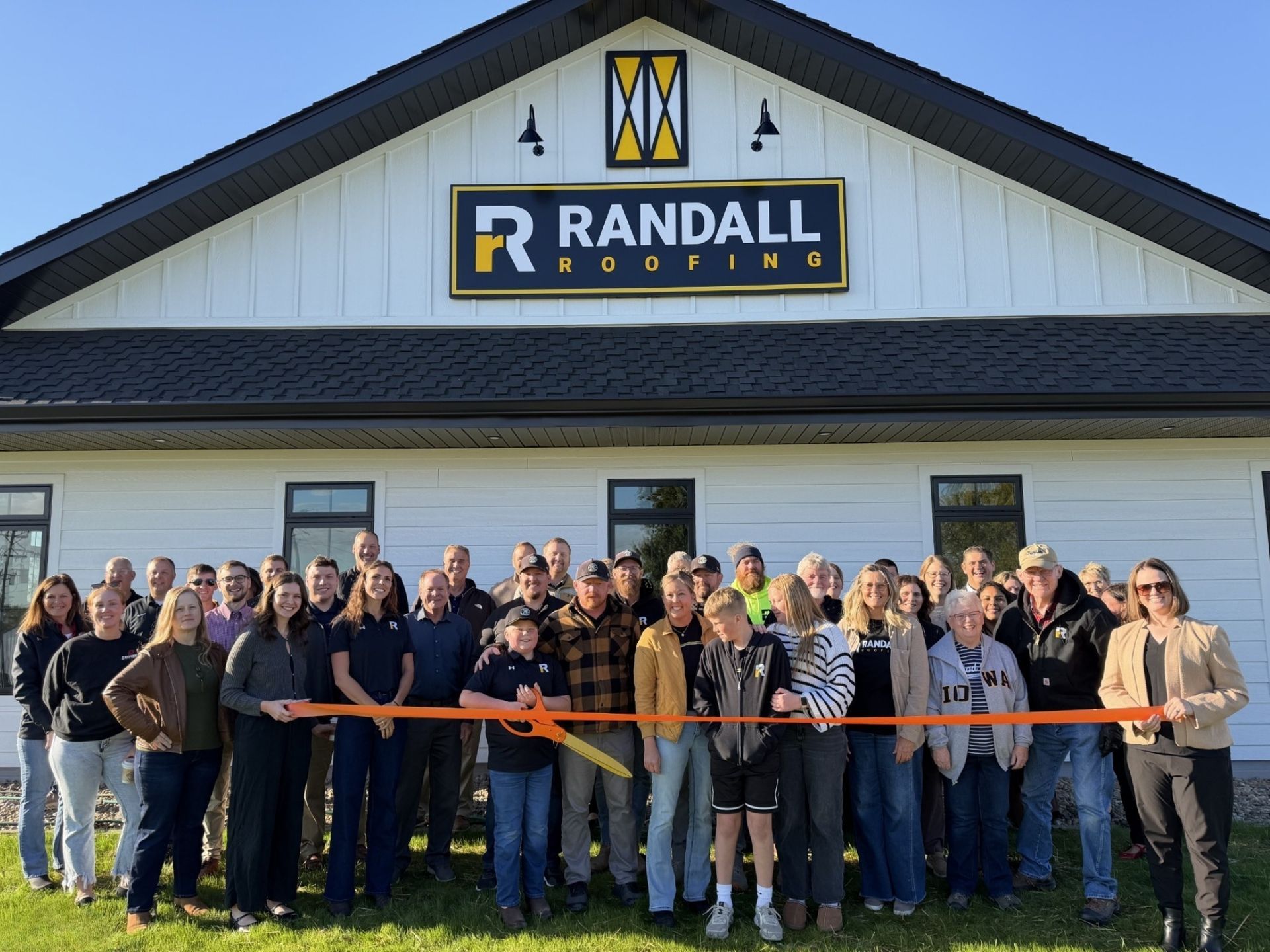 A large group gathers for a ribbon-cutting ceremony in front of a white building with a Randall Roofing sign.