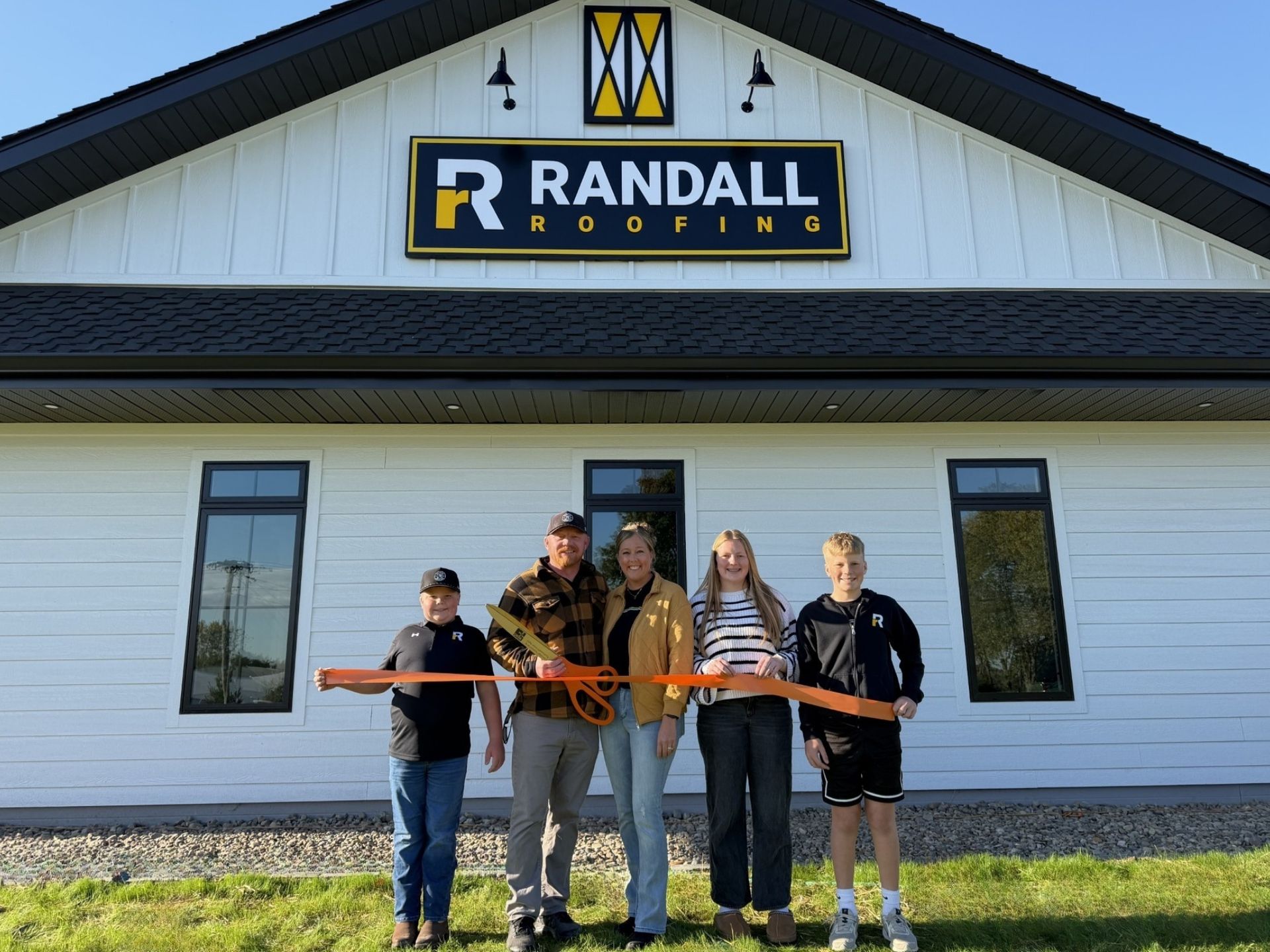 A family stands in front of a Randall Roofing building holding ceremonial scissors to a ribbon during a grand opening.