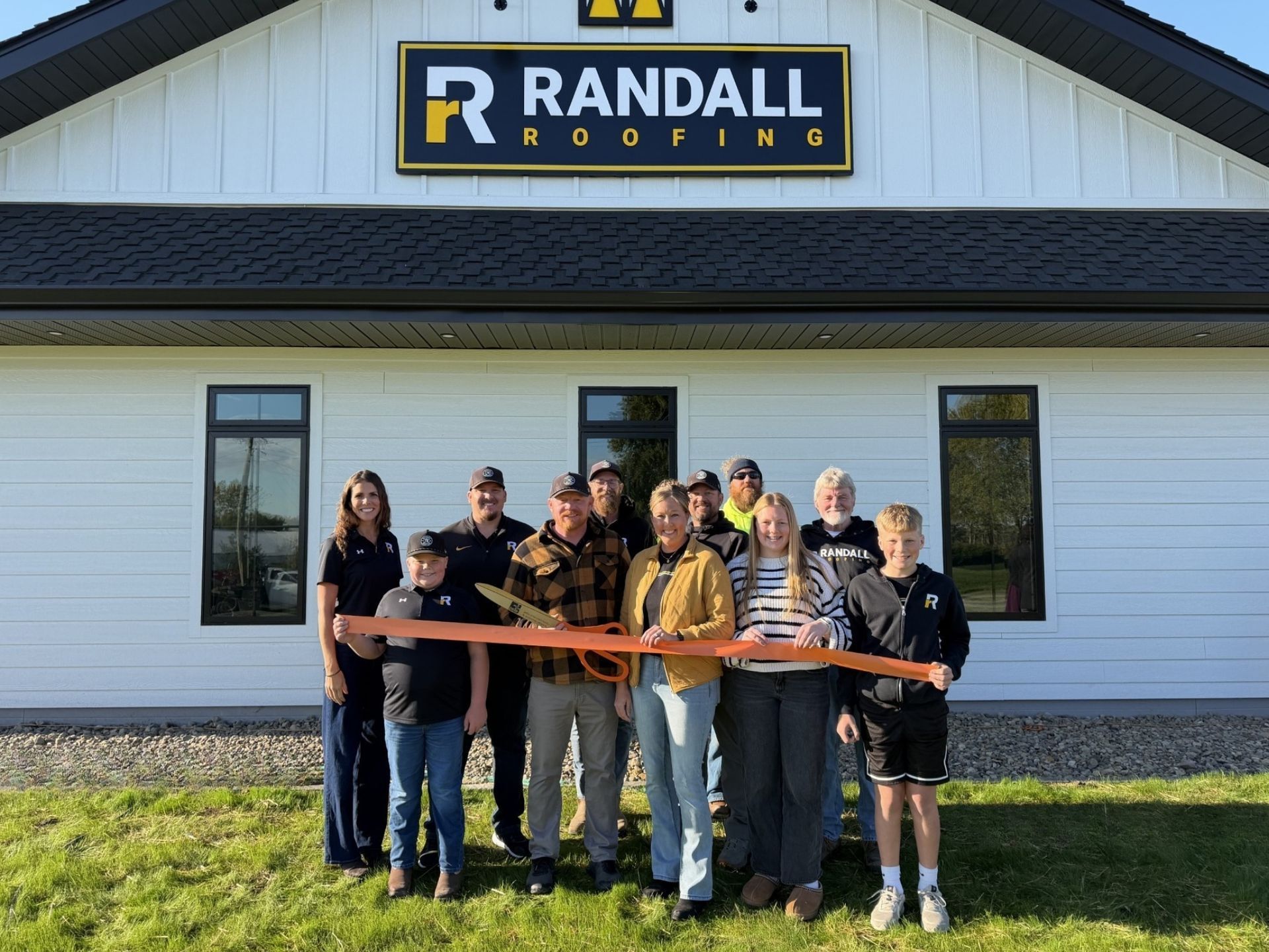 A group of people holding a large ribbon at a ribbon-cutting ceremony in front of a Randall Roofing office building.