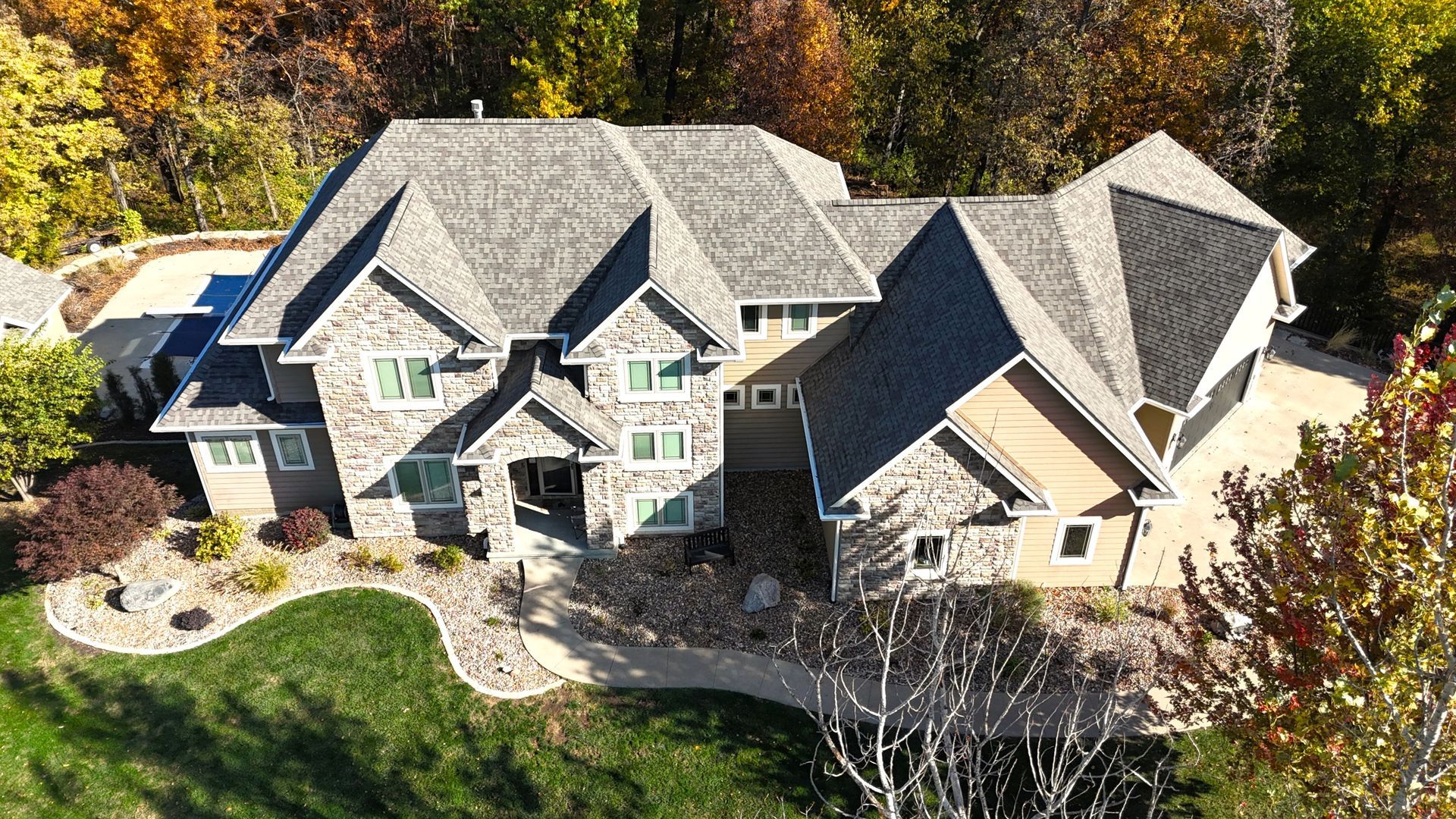 Aerial view of a large stone and stucco house with a shingled roof, surrounded by trees and a landscaped yard.