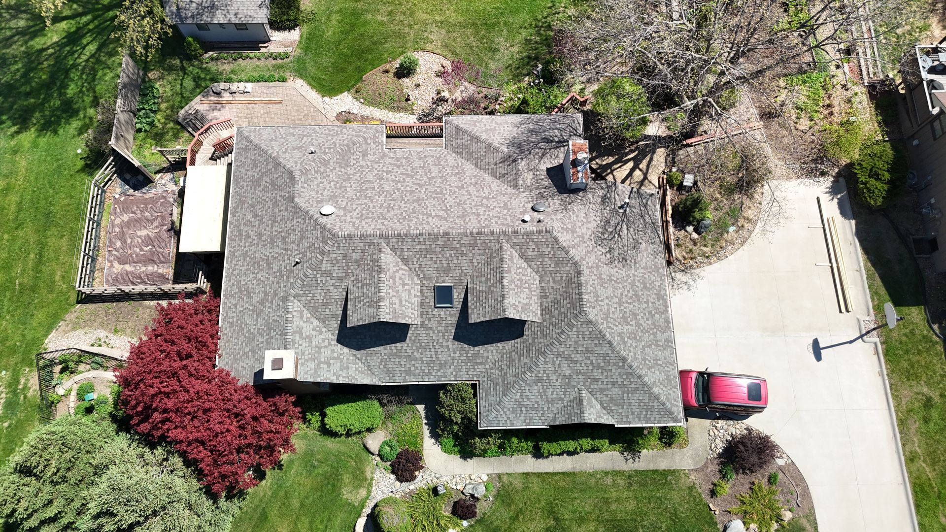 An aerial view of a single-story house with a complex shingled roof, surrounded by green lawns, trees, and a driveway.