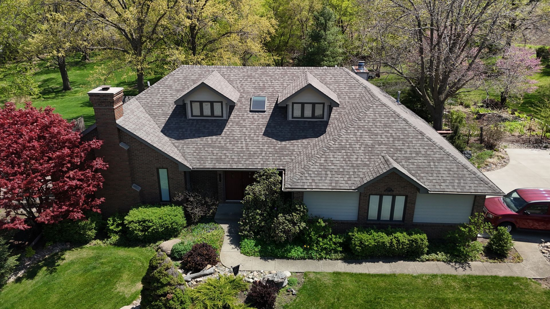 An aerial view of a suburban house with a gray shingled roof, two dormers, a brick chimney, and a red car in the driveway.