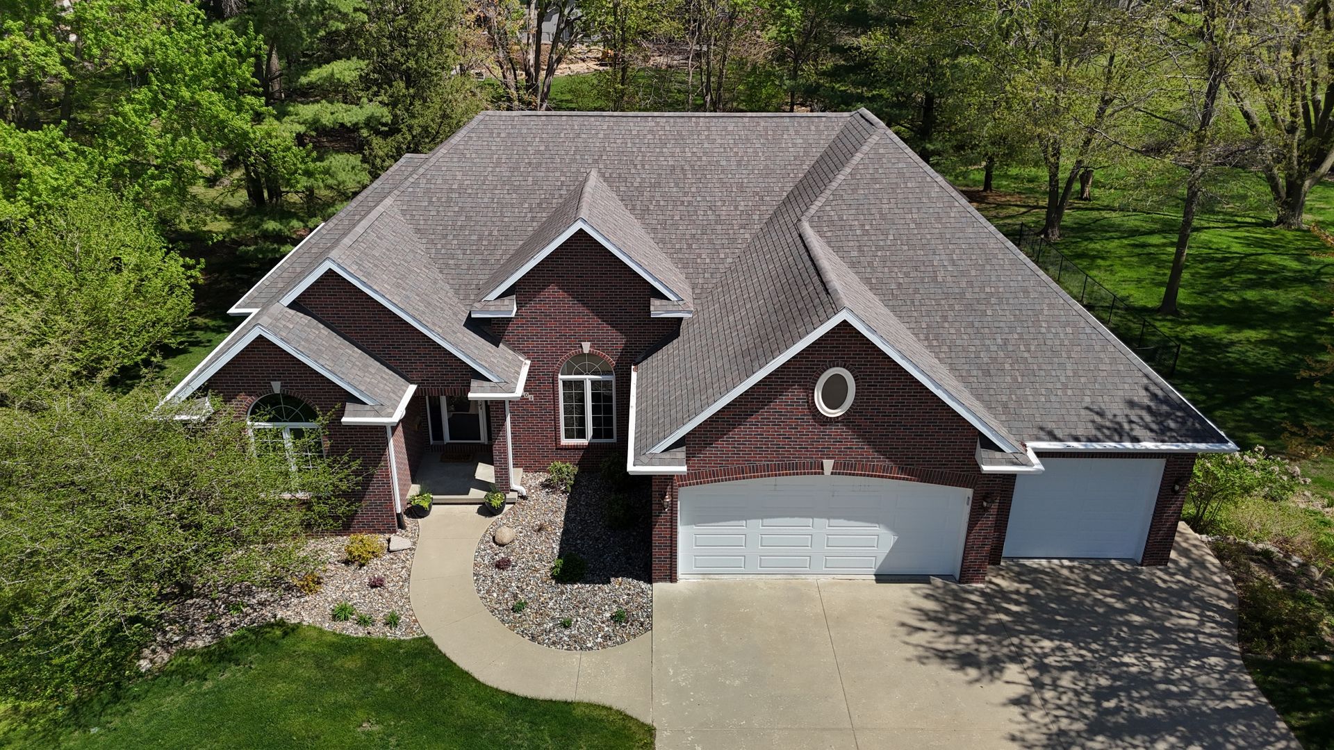 An aerial view of a brown brick house with a dark gray shingled roof, a paved driveway, and lush green trees.