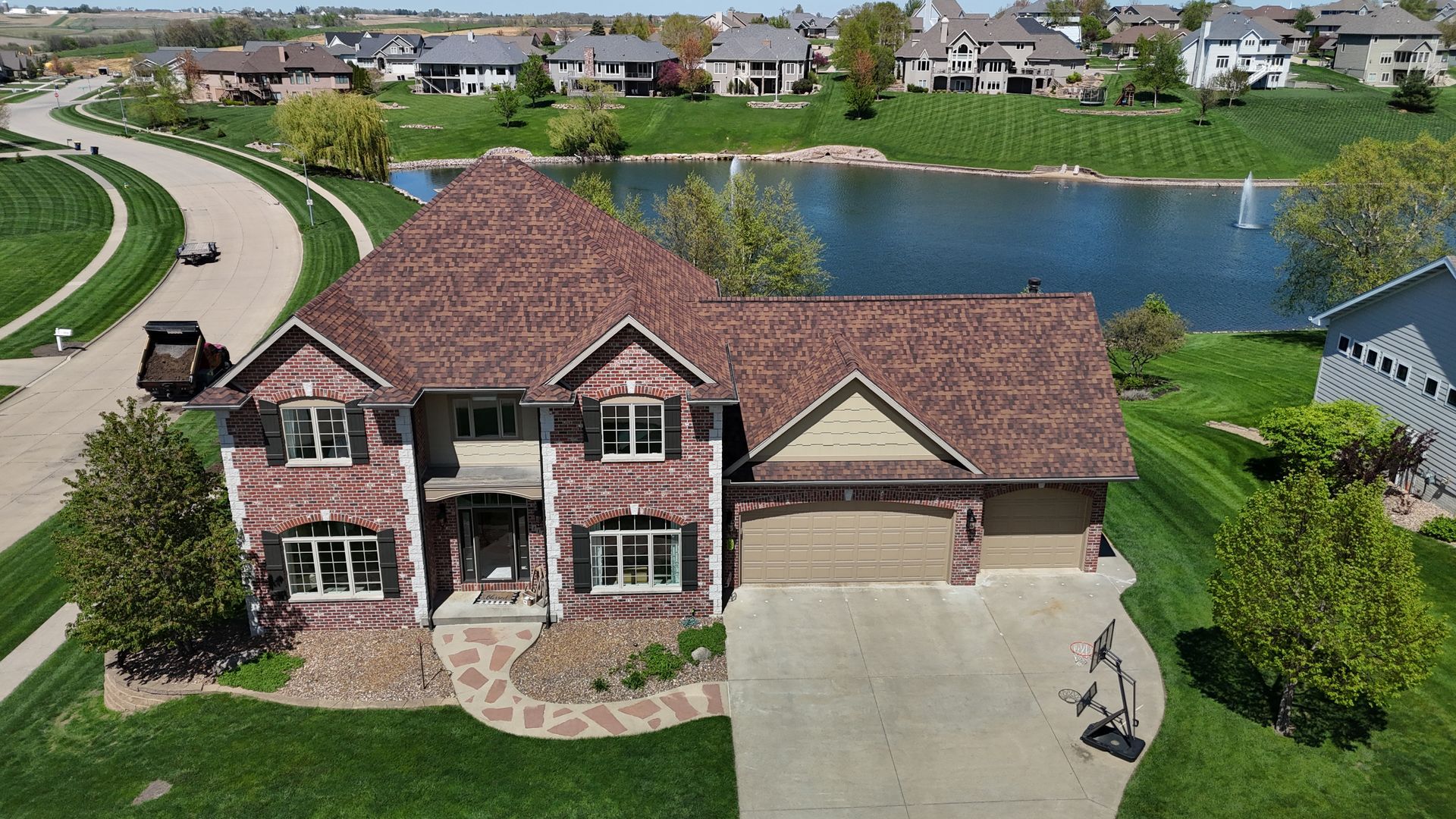 An aerial view of a two-story brick house with a dark shingled roof, a paved driveway, and a backyard facing a pond.