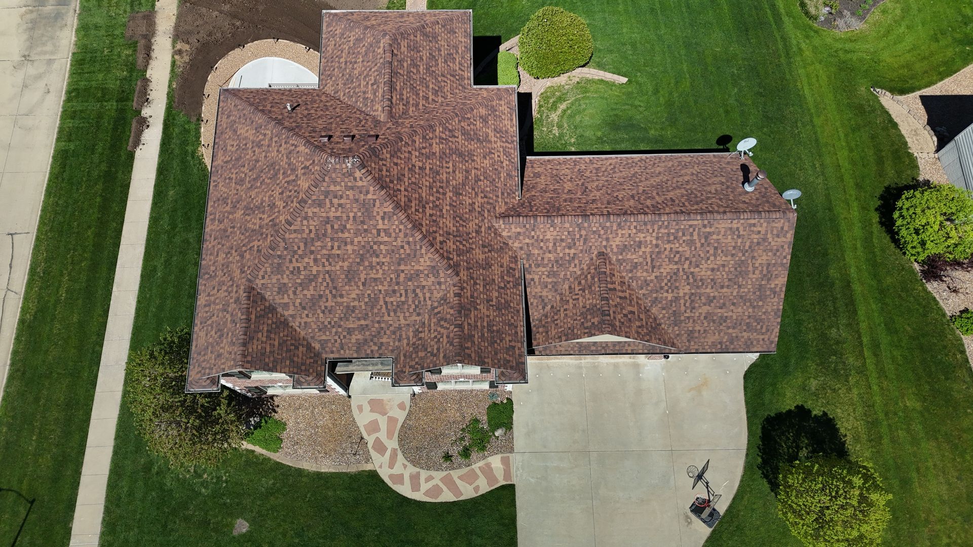 Aerial view of a residential house with a brown shingled roof, surrounding lawn, driveway, and stone walkway.