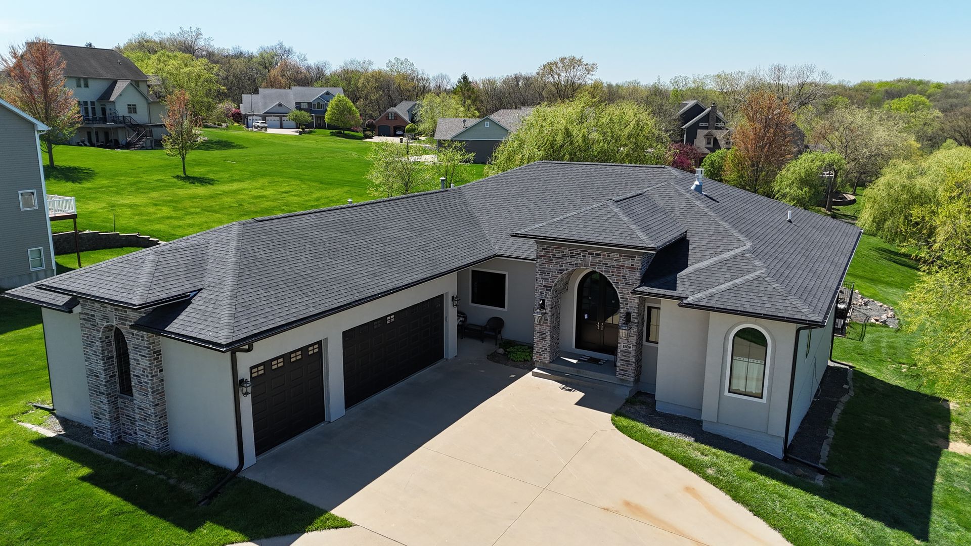 Aerial view of a gray stucco ranch-style house with a dark shingled roof, stone accents, and a driveway in a suburban area.