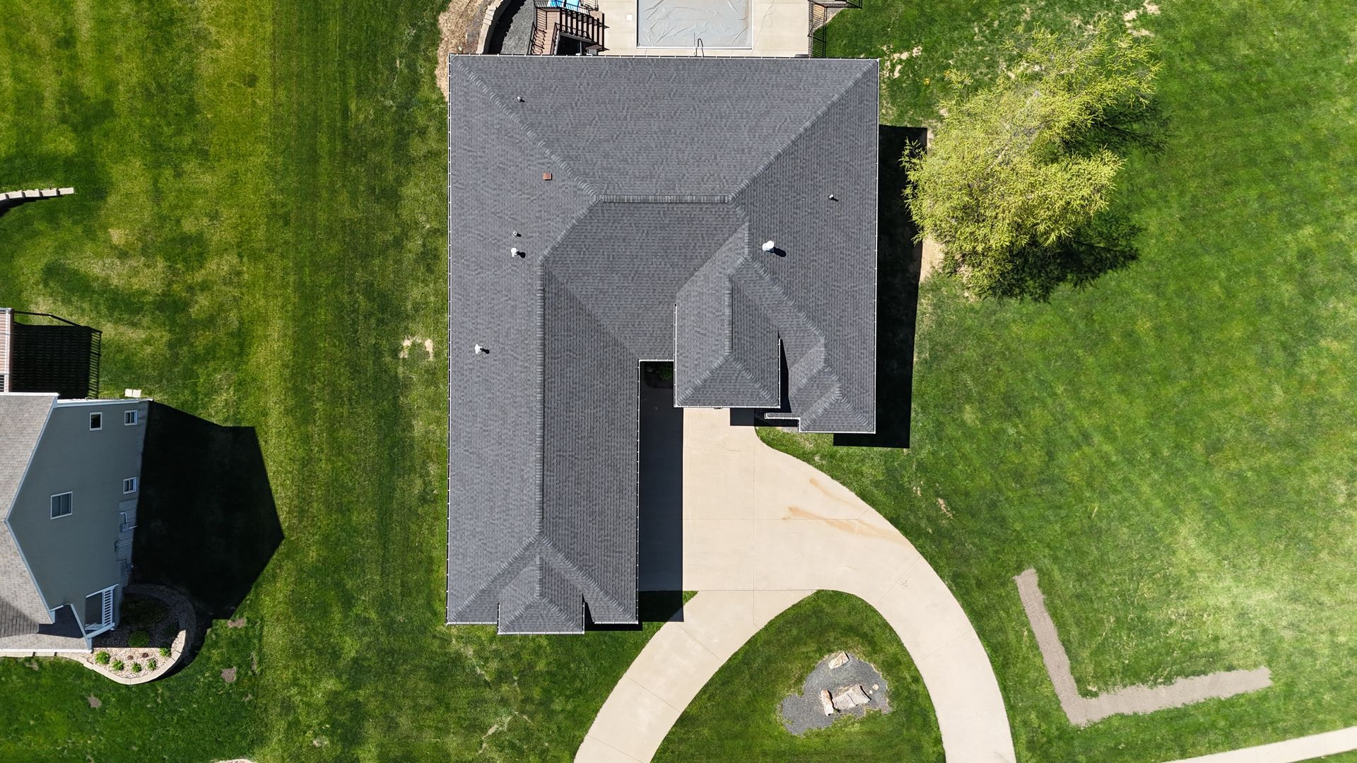An aerial view of a gray-roofed L-shaped house surrounded by a green lawn and a curved concrete driveway.