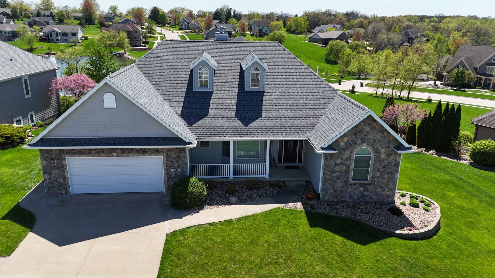 An aerial view of a suburban home with a gray shingled roof, stone accents, a white garage door, and a green lawn.