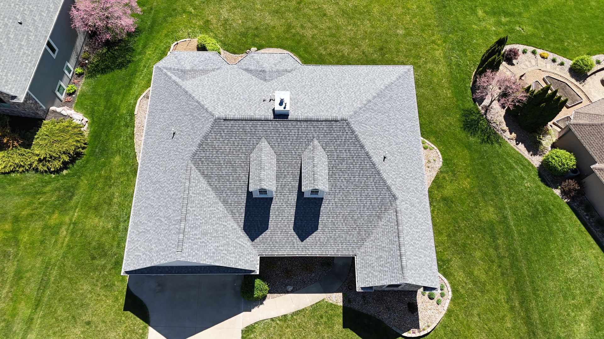 Aerial view of a residential house with a gray shingled roof, two dormers, and a chimney, surrounded by green lawns.