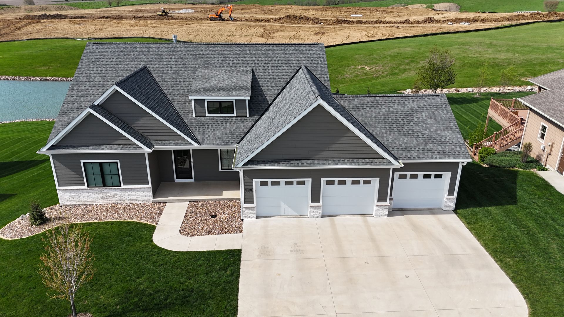 An aerial view of a dark grey house with a white stone base, three-car garage, and grey roof, situated on a grassy lot.