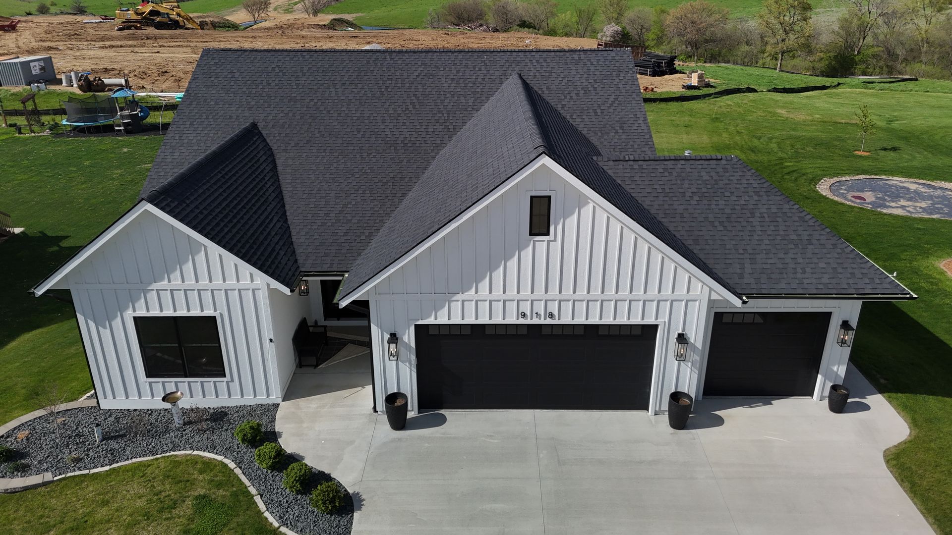 Aerial view of a white, modern farmhouse with a dark grey roof and three garage doors, set on a green, landscaped lawn.