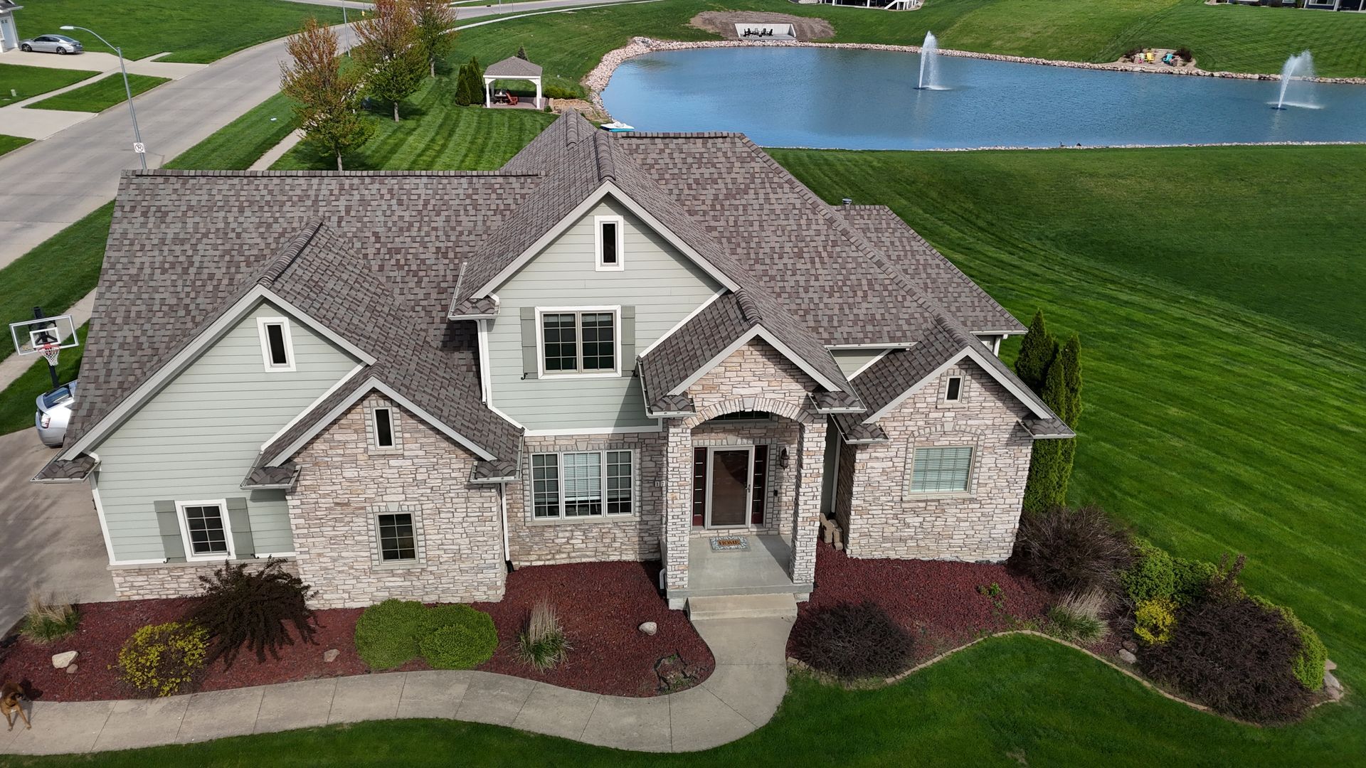An aerial view of a two-story house with stone and gray siding, featuring a paved walkway and a nearby pond with fountains.