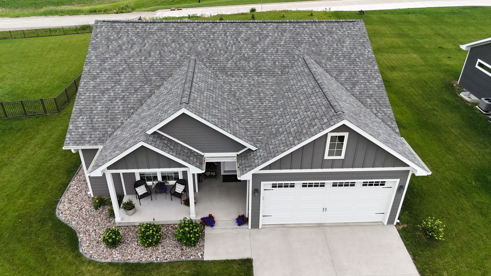 An aerial view of a gray suburban house with a white garage, covered front porch, and surrounding green lawn.
