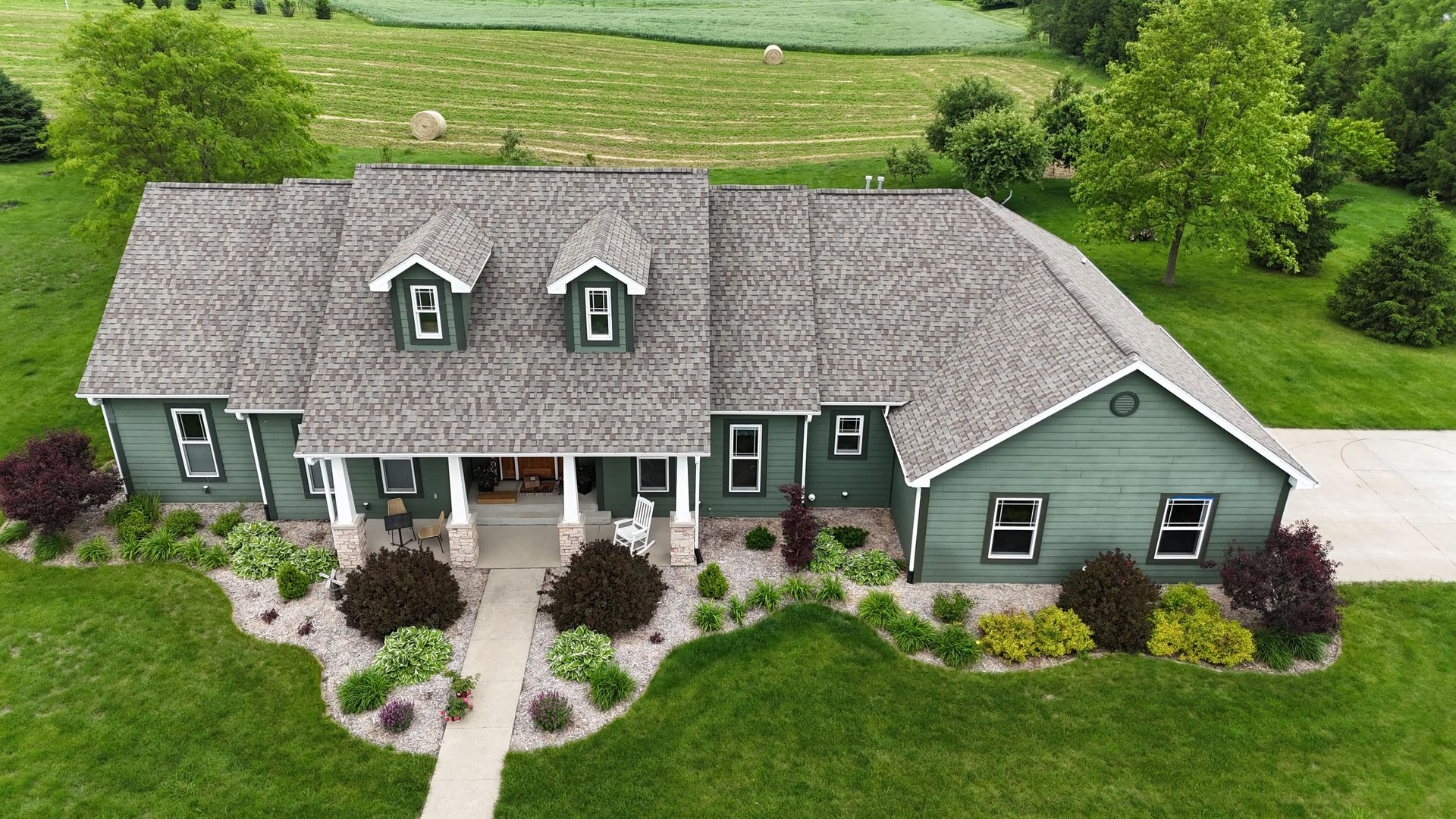 An aerial view of a sage green, single-story house with a gray shingled roof, surrounded by lawn and landscaping.
