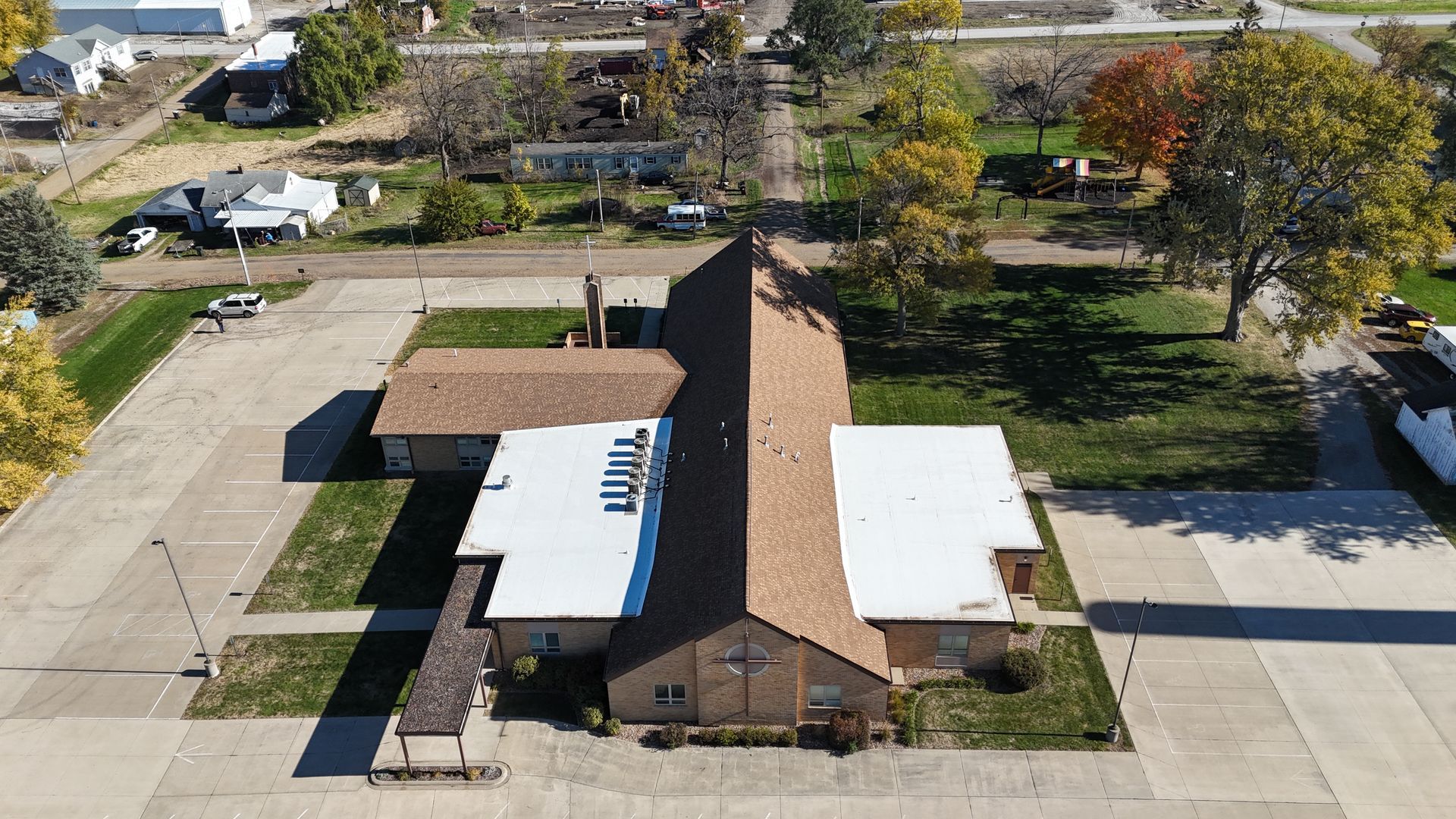 Aerial view of a church building with a brown roof and white additions surrounded by a paved parking lot and trees.