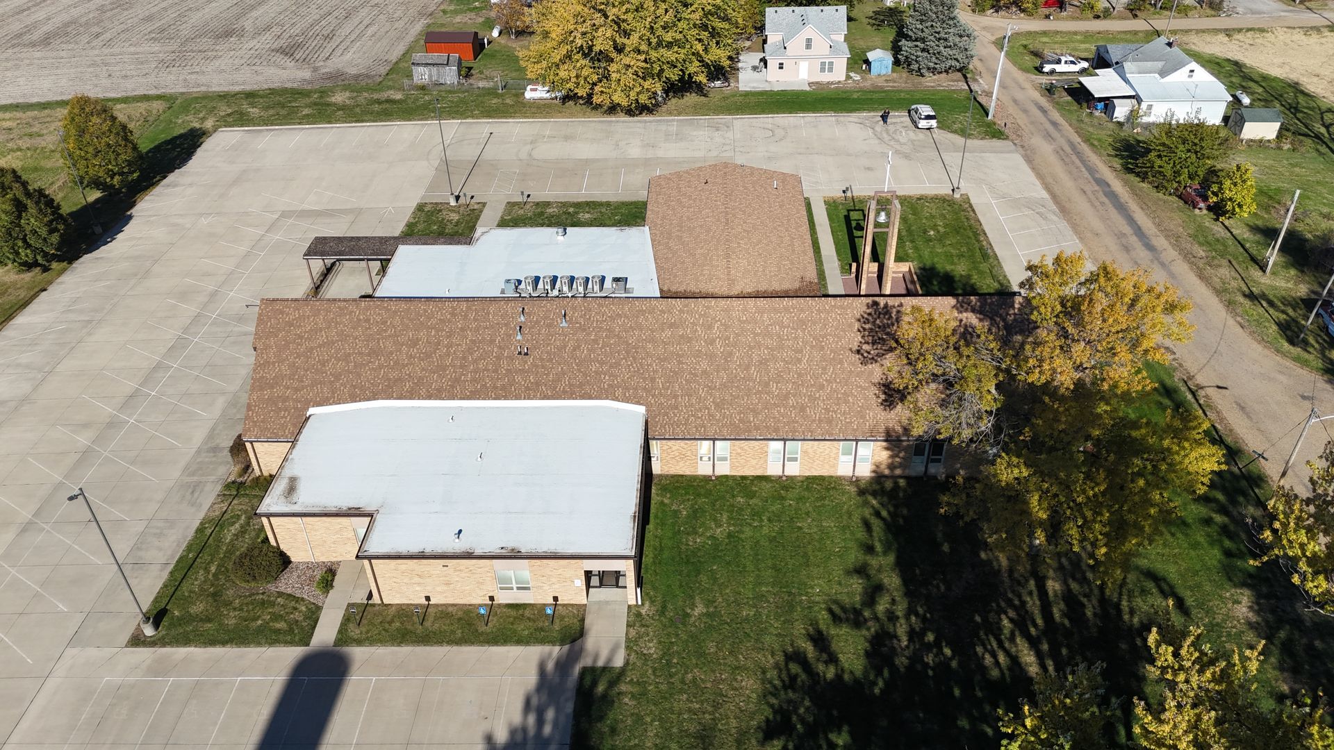 Aerial view of a building with a large parking lot, surrounded by trees and neighboring houses in a rural setting.
