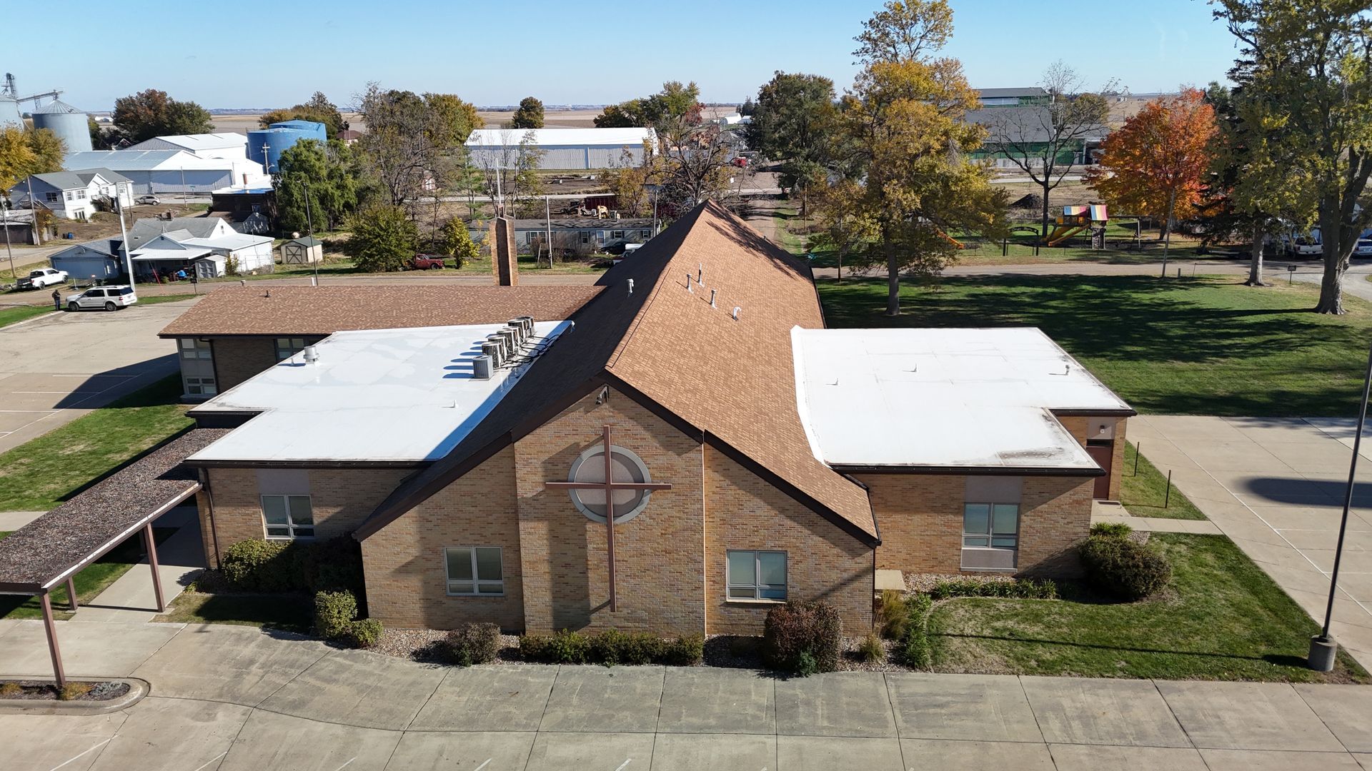 Aerial view of a brick church with a large, centered wooden cross on the front exterior, surrounded by trees and pavement.