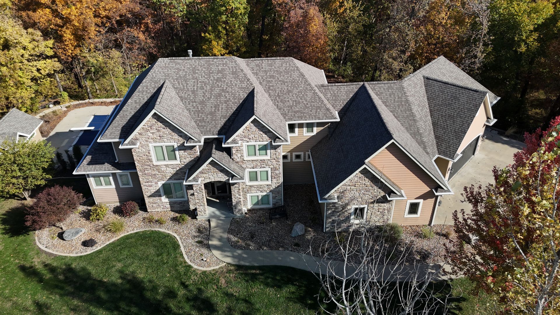 An aerial view of a multi-story stone and beige home with a grey roof surrounded by autumn trees and a manicured lawn.