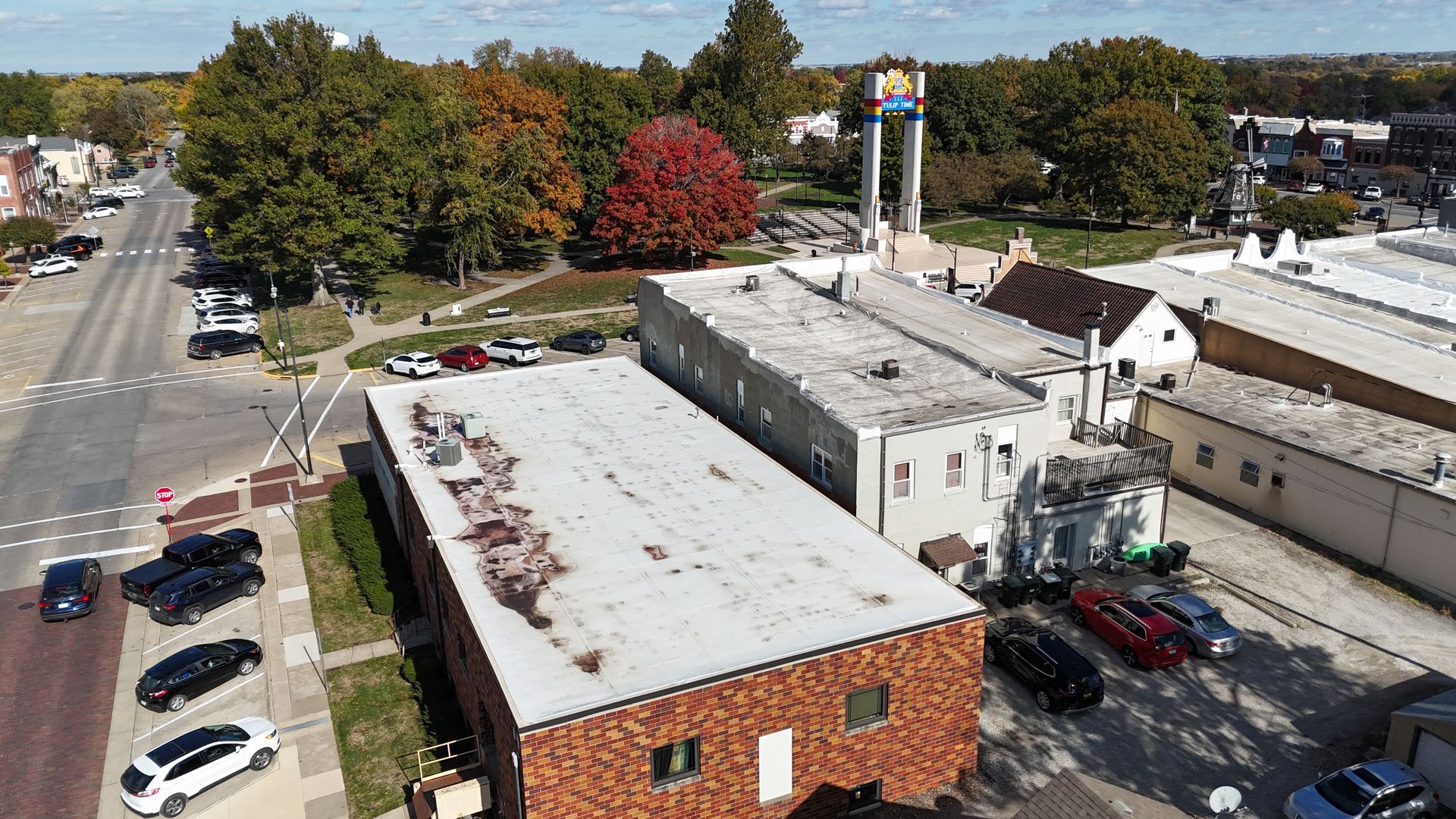Aerial view of a small town street, buildings with flat roofs, a park with a tall monument, and trees in autumn colors.