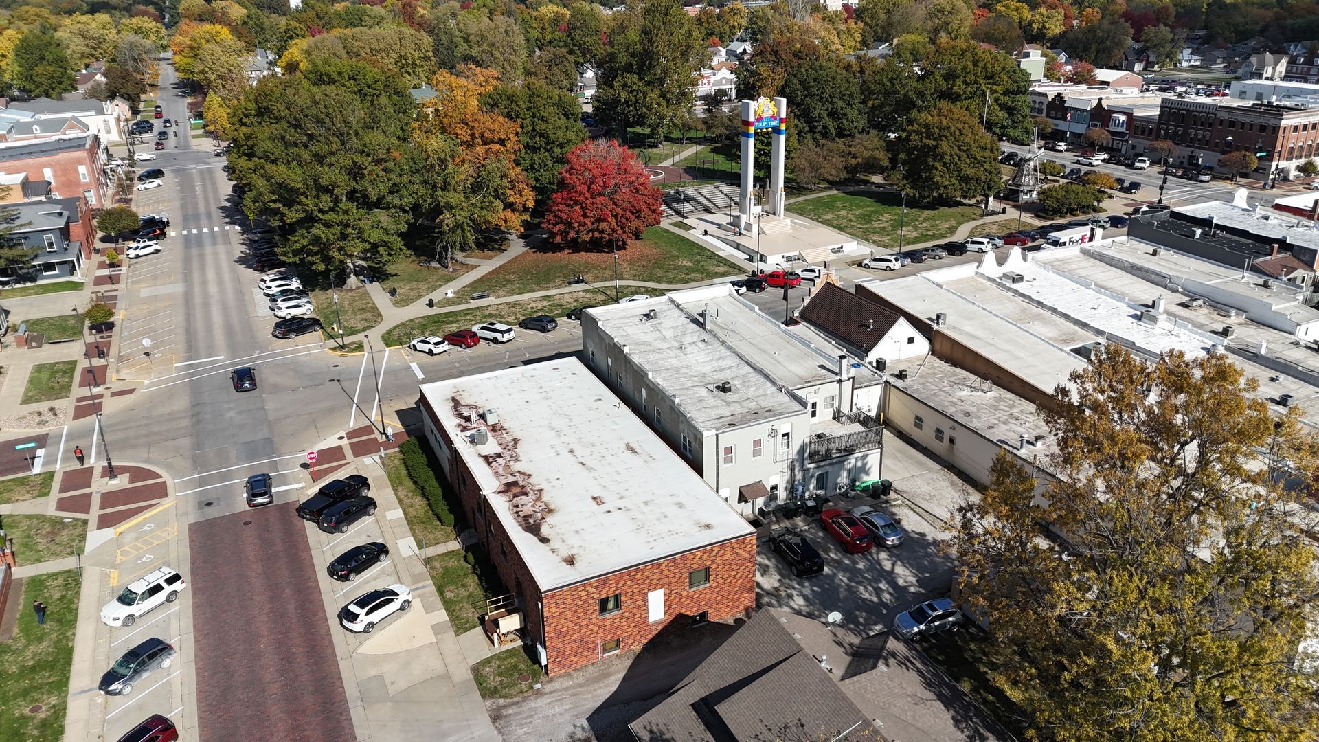 Aerial view of a small town street, buildings with flat roofs, a green park with a monument, and parked cars in autumn.
