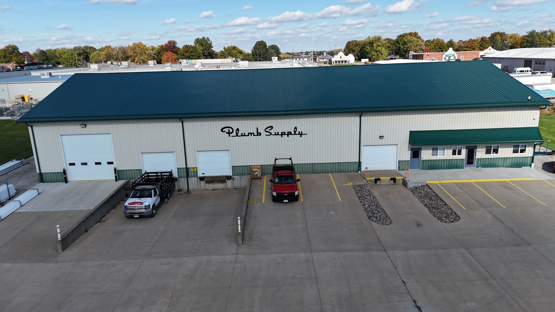 An aerial view of a light-colored warehouse building with a green roof, featuring a loading dock area and two parked vehicles.