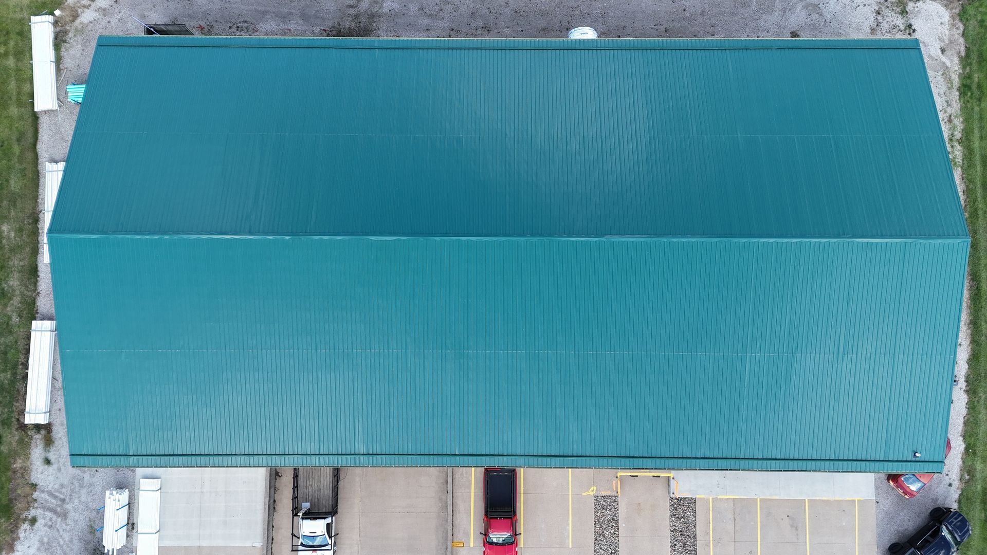An aerial view of a rectangular building with a green metal roof, situated above a parking lot with a few vehicles.