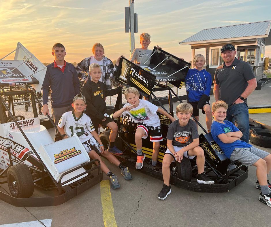 Group of people posing by two small race cars on a paved track at sunset.