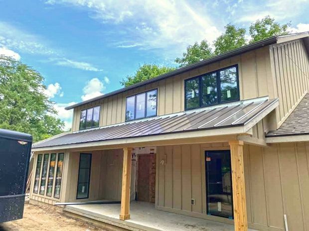 A two-story tan home with board-and-batten siding, a metal porch roof, and black-framed windows under a blue sky.